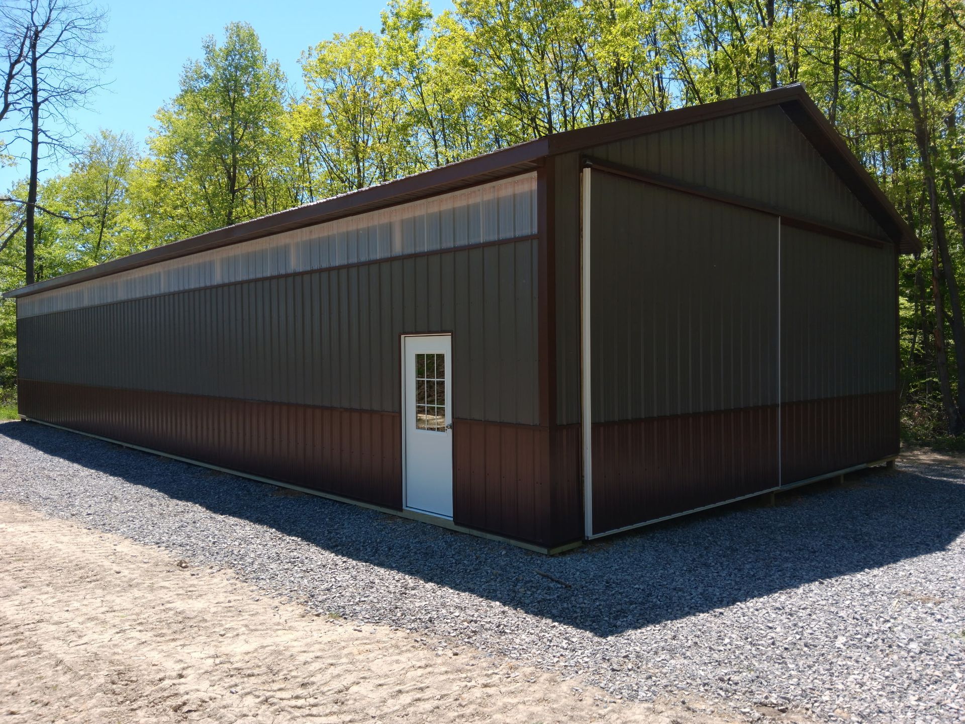 Dark metal shed with a white door on a gravel lot, with trees and blue sky behind it