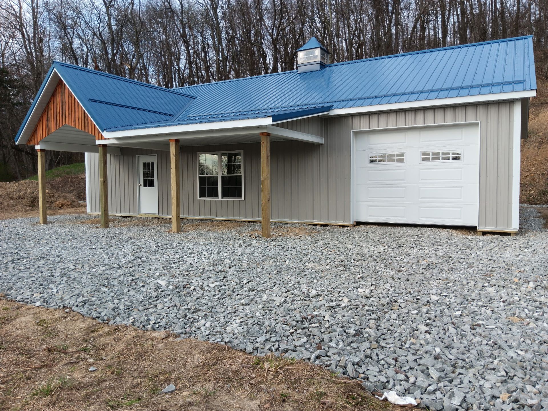 Small gray house with blue metal roof, front porch, and attached white garage on gravel.