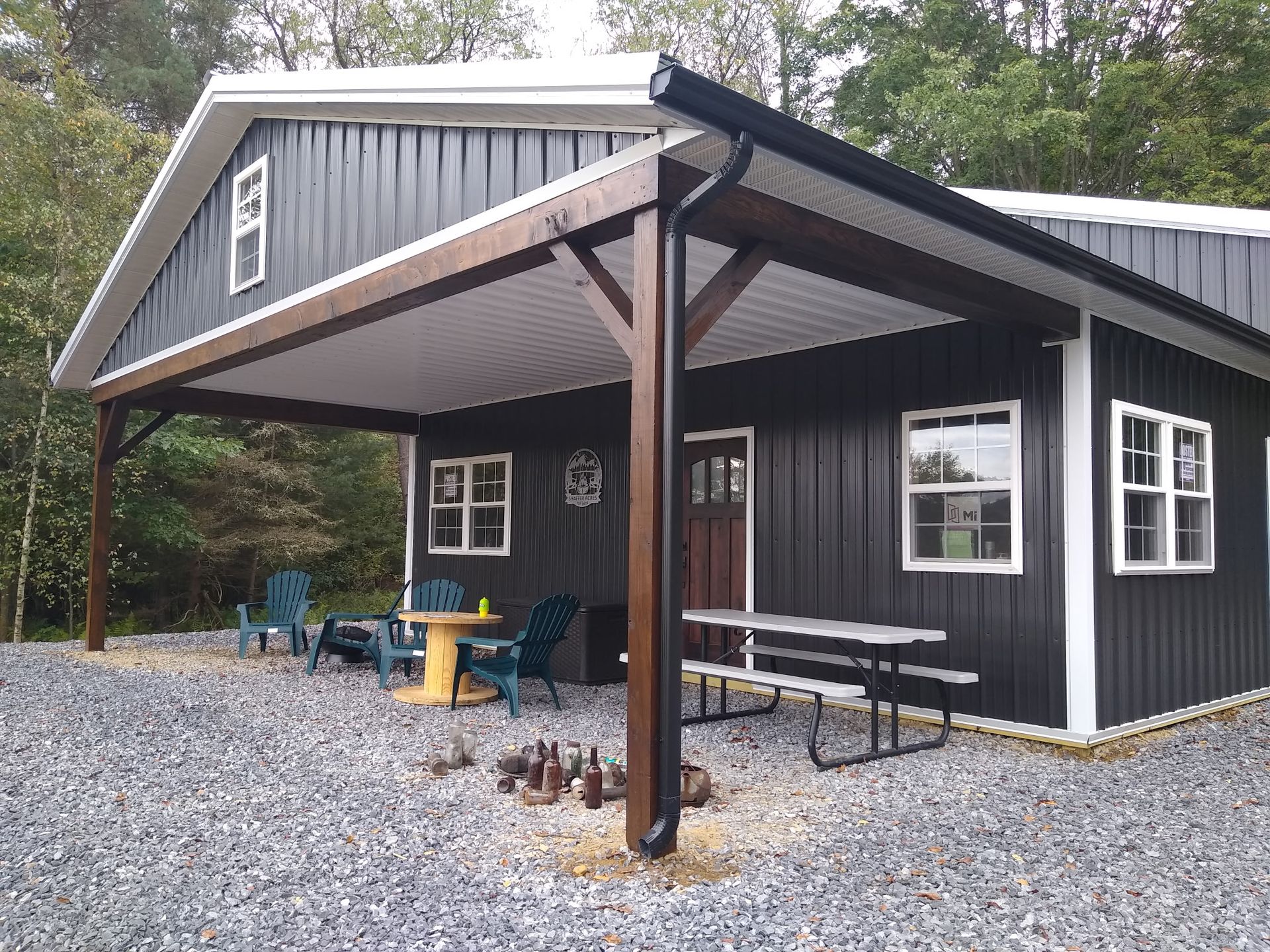 Black cabin-style pavilion with covered porch, picnic tables, and gravel clearing in a wooded area