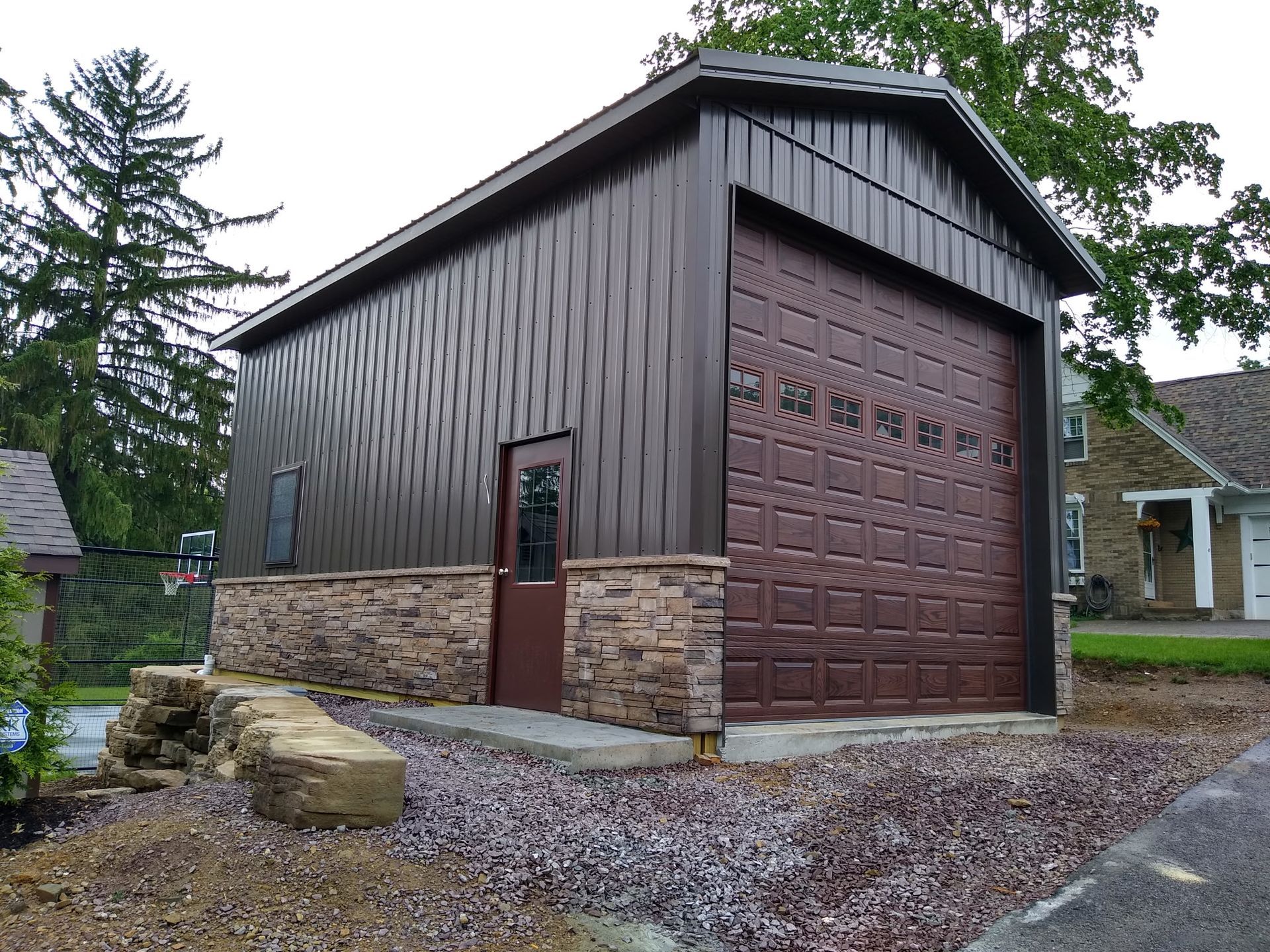 Brown barn-style garage with stone base and large wooden door on a gravel driveway