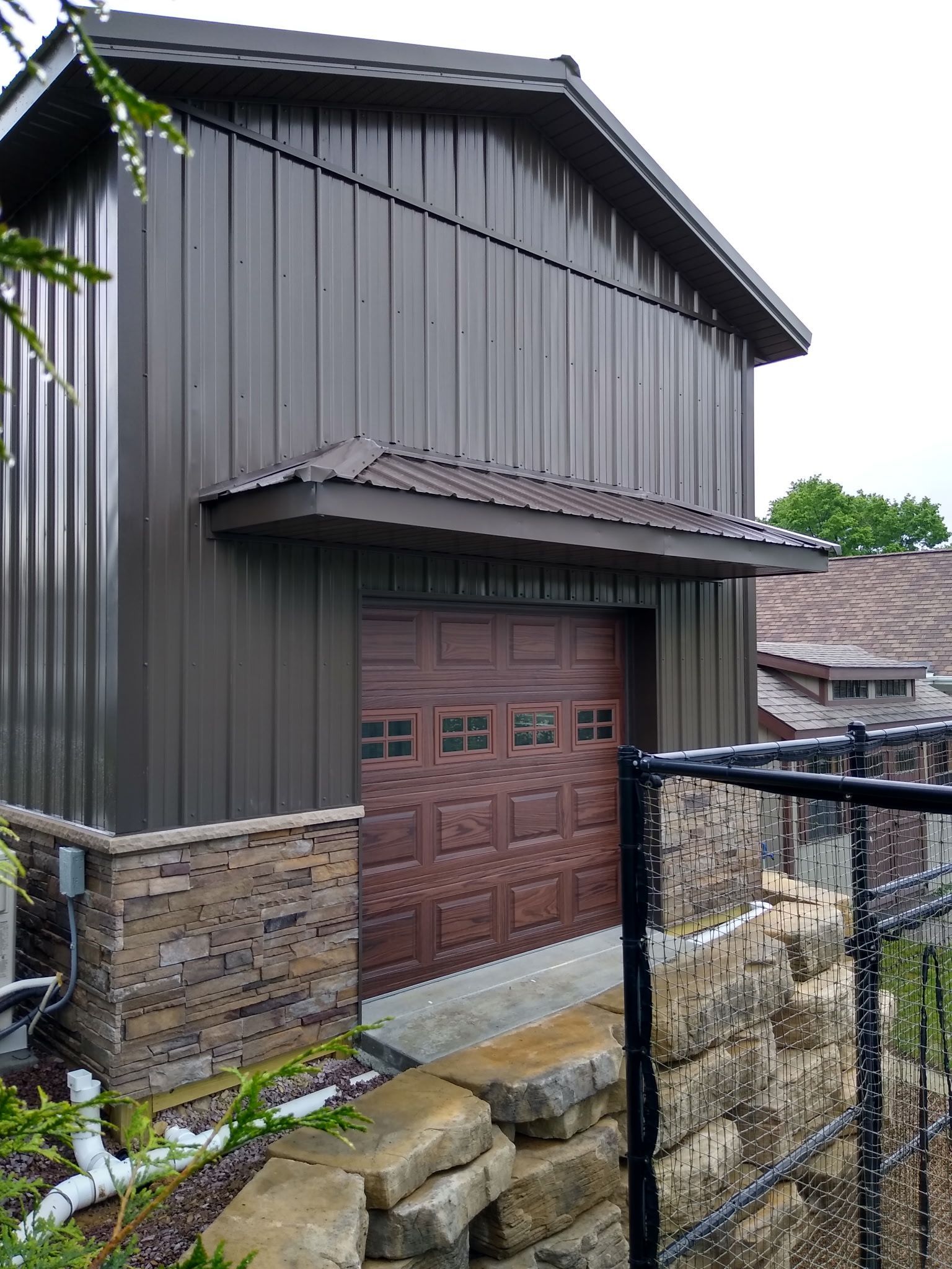 Gray barn-like garage with a brown wooden door, stone base, and chain-link fence in front