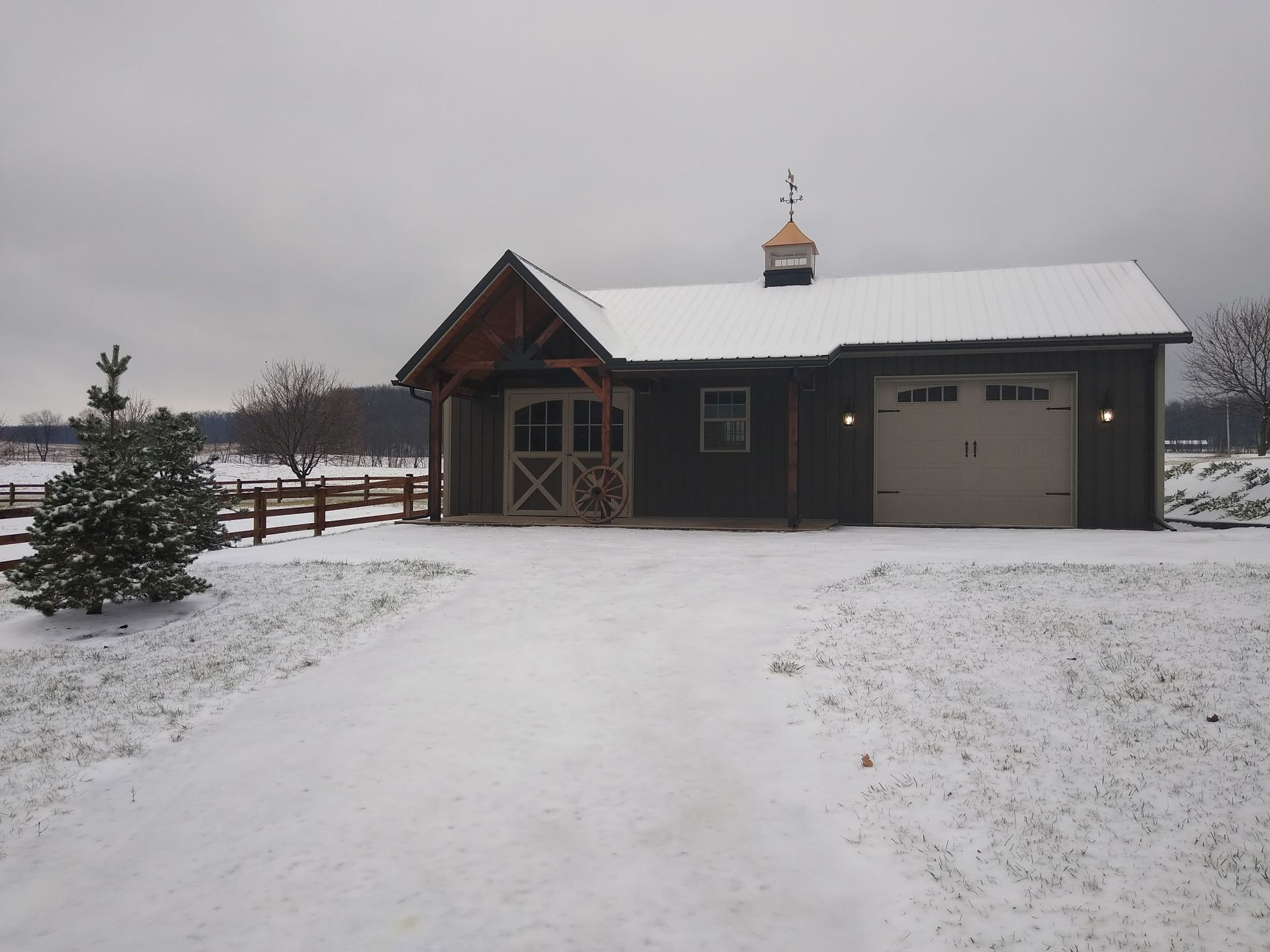 Snow-covered driveway leading to a dark garage and small cabin under an overcast winter sky