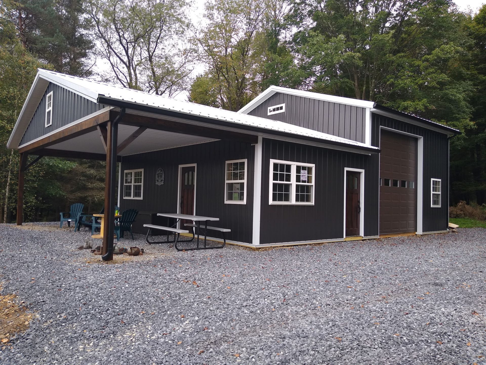 Black cabin with a covered porch and picnic table on a gravel lot, surrounded by trees.