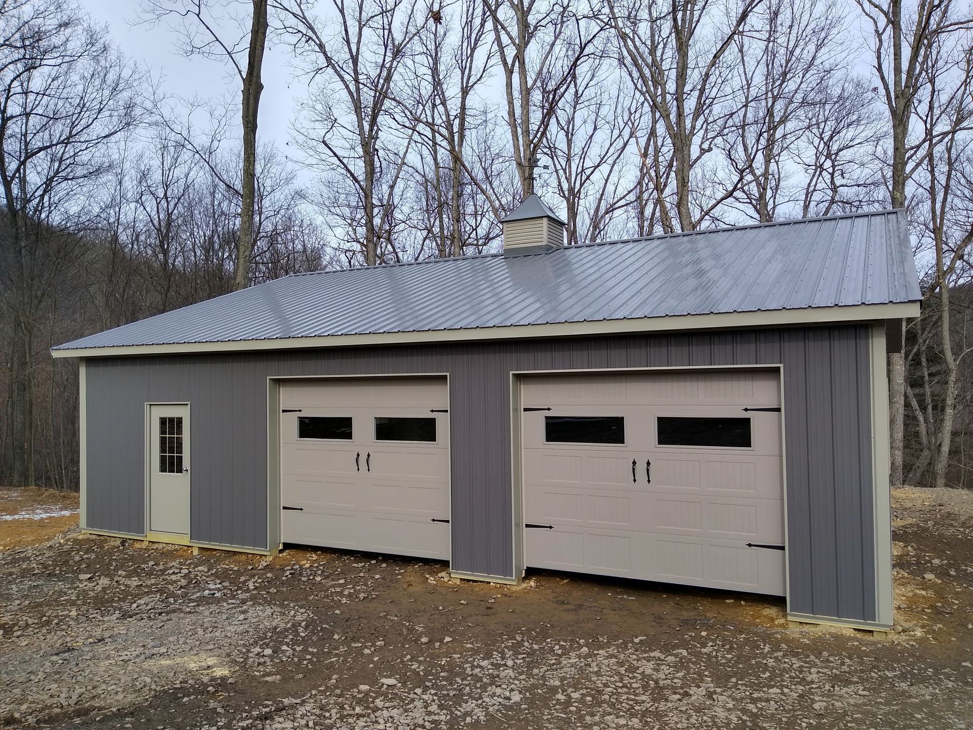 Gray two-car garage with side door and metal roof in a wooded outdoor setting