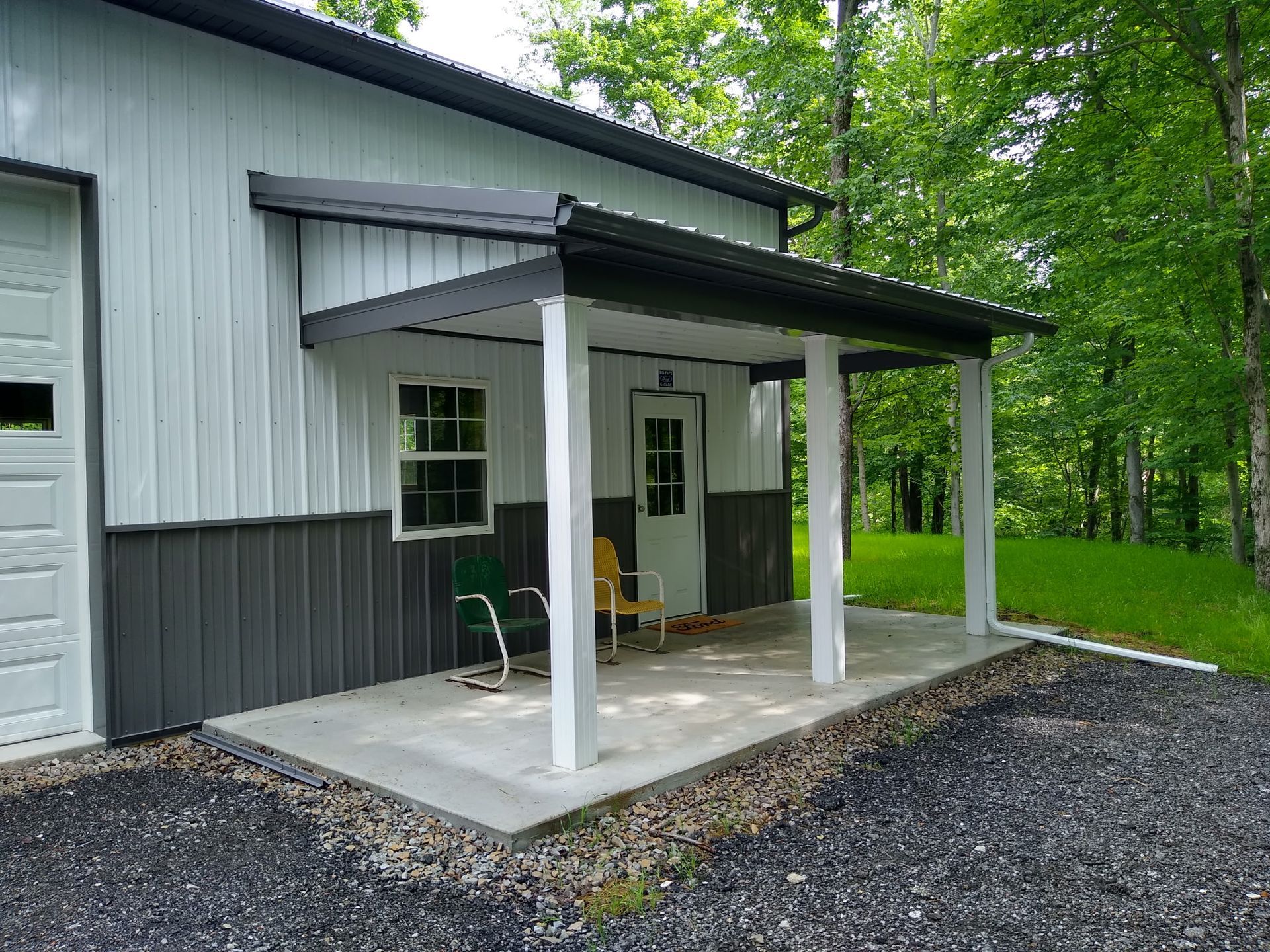Small gray house with a covered front porch, white columns, and gravel driveway surrounded by trees