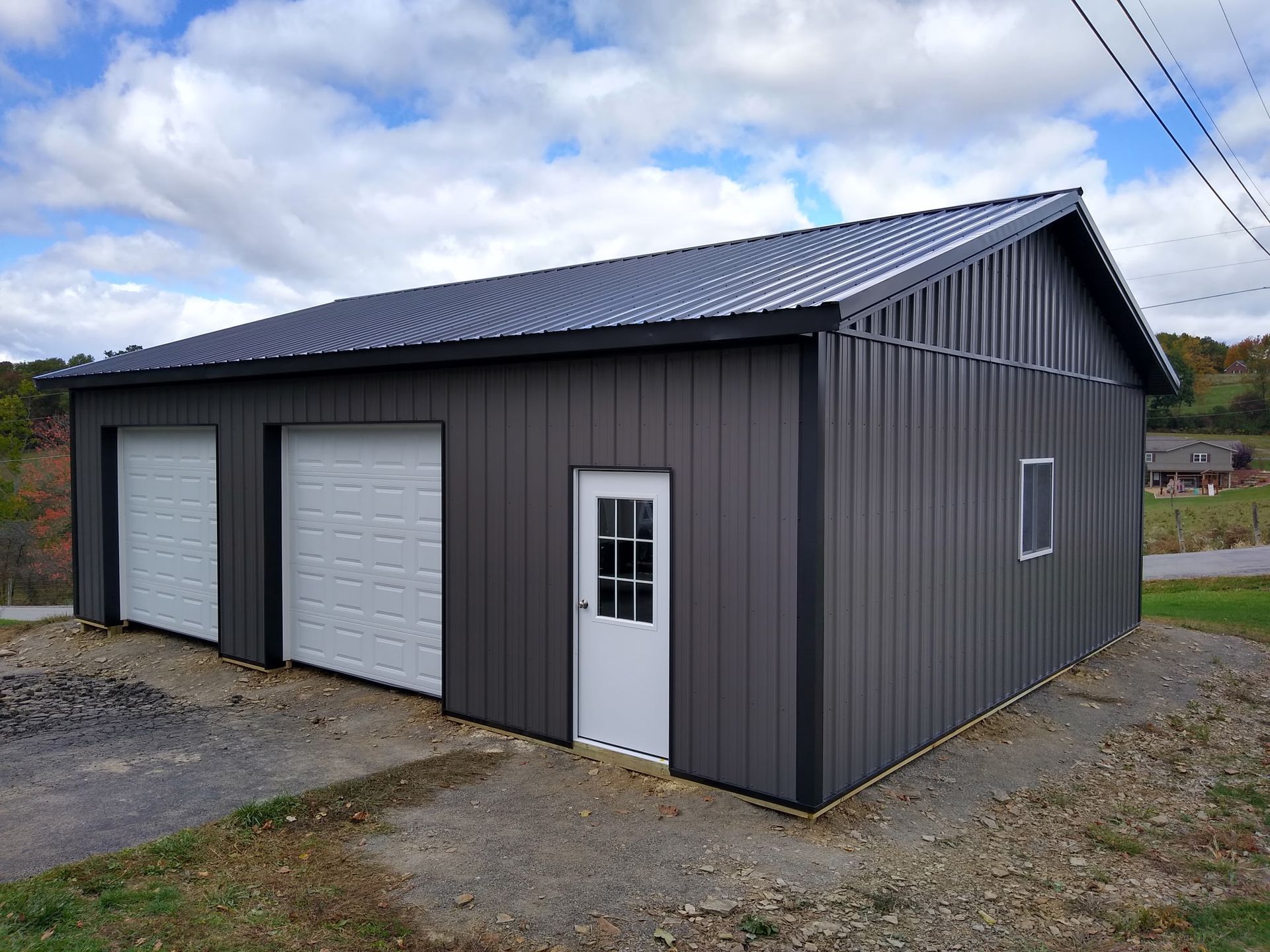Gray metal garage with two white overhead doors and a side entry door on a gravel lot under a cloudy sky