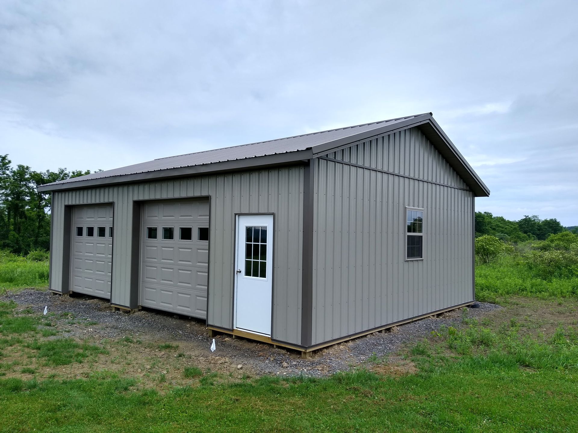 Gray metal garage building with two roll-up doors and a side entry door in a grassy field