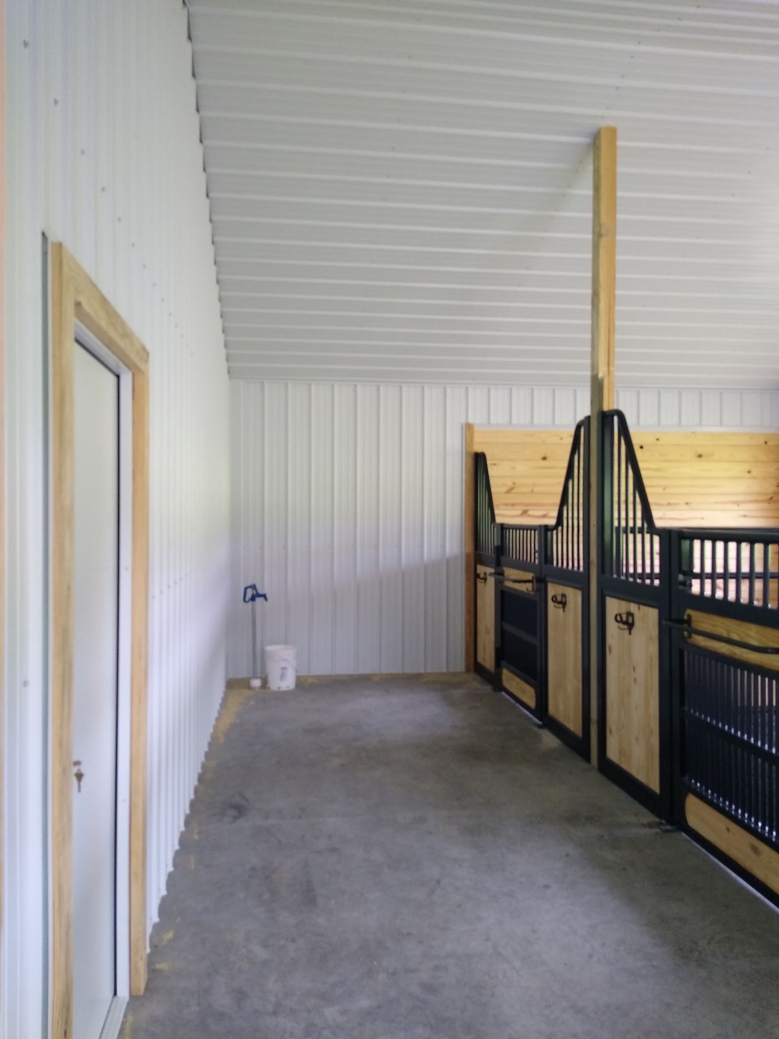Concrete barn aisle with white paneled walls, black metal stalls, and a closed wooden door.