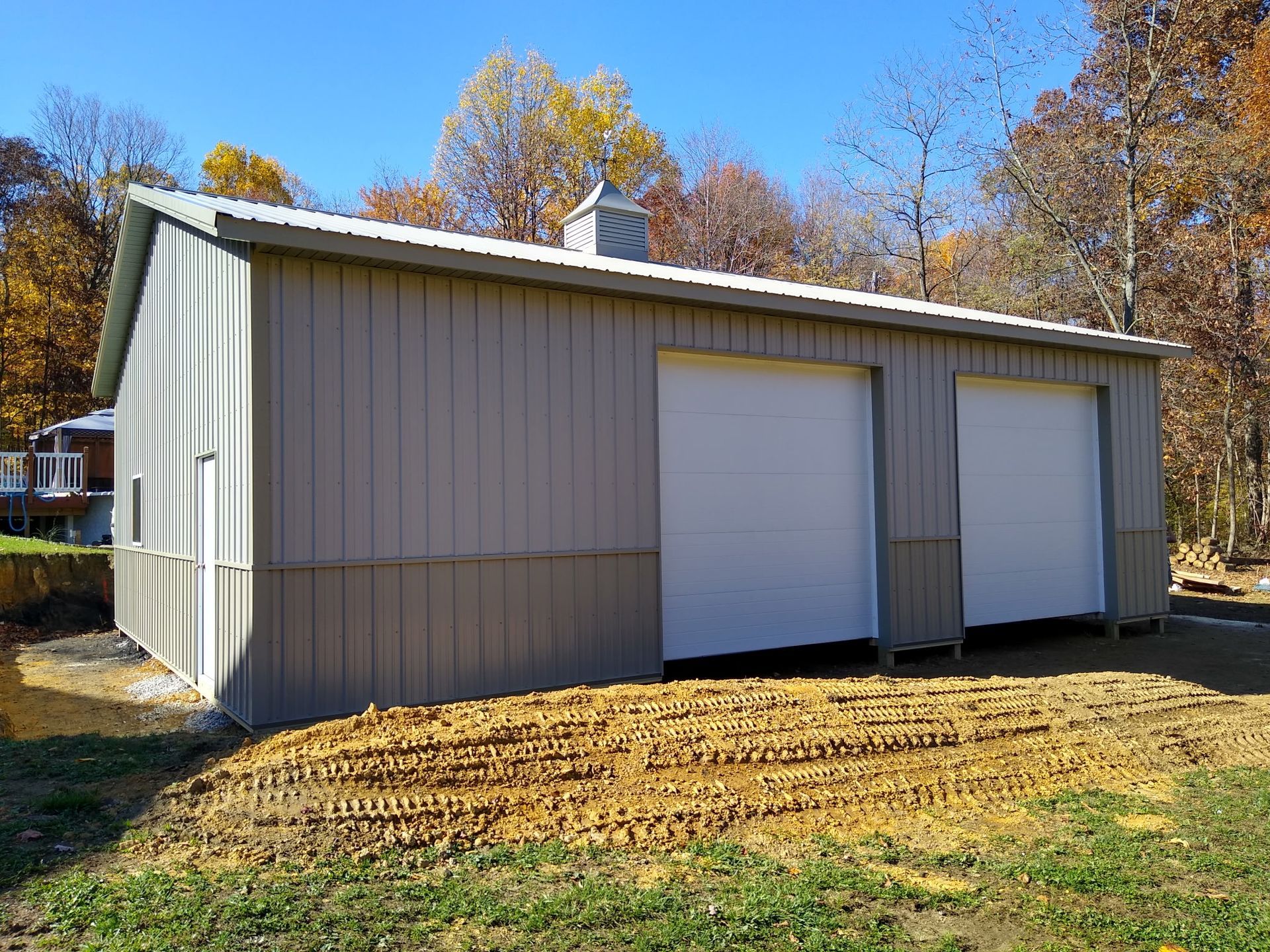 Small beige metal shed with two white garage doors on a sunny, leaf-covered lot