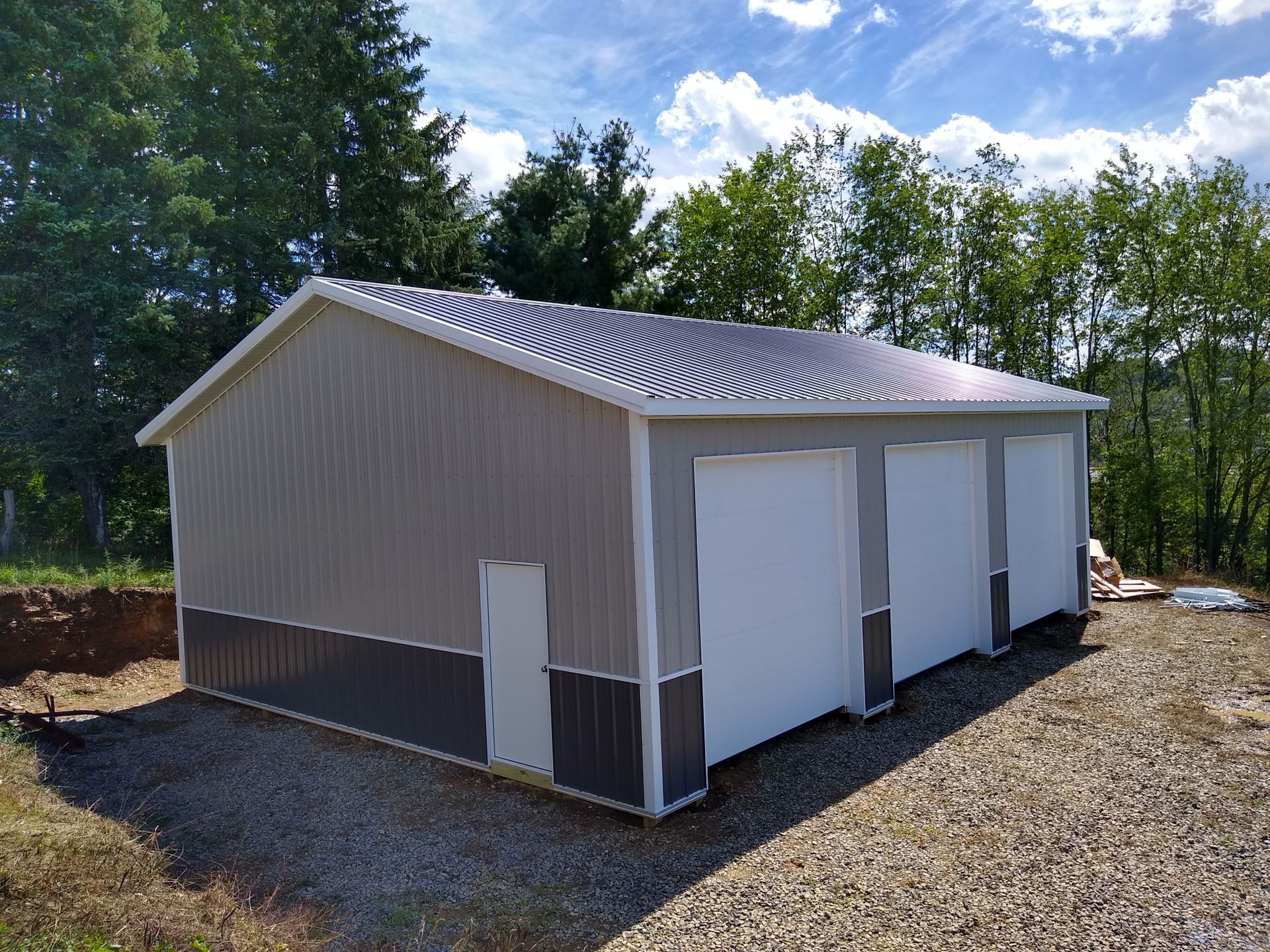 Small beige metal garage or storage building with three white roll-up doors on a gravel lot, surrounded by trees.