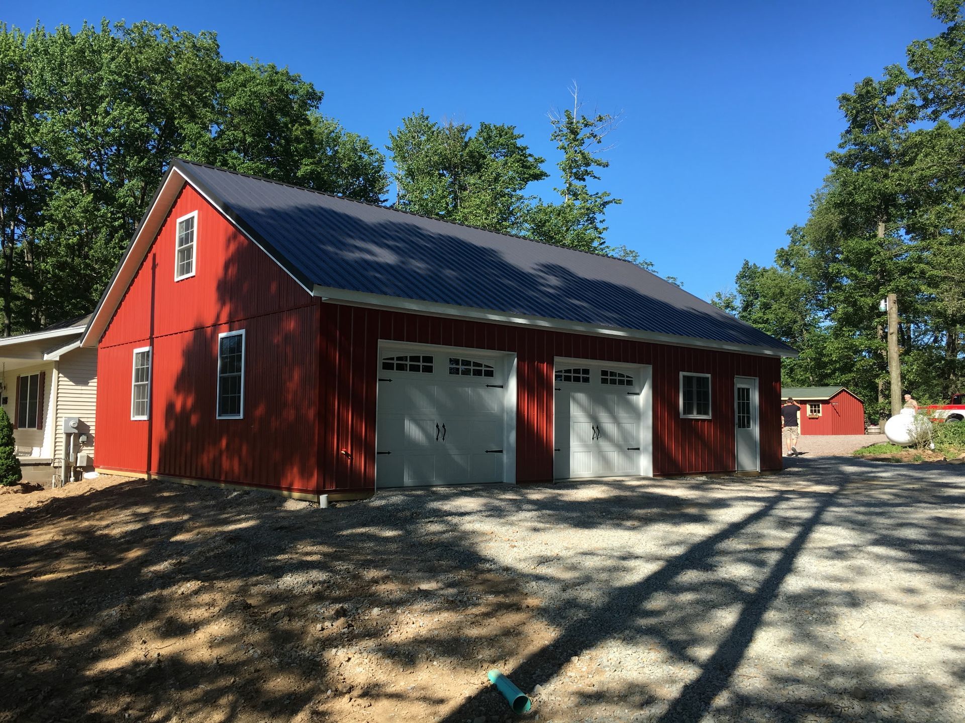 Red barn-style garage with two white doors and a black roof in a sunny wooded yard