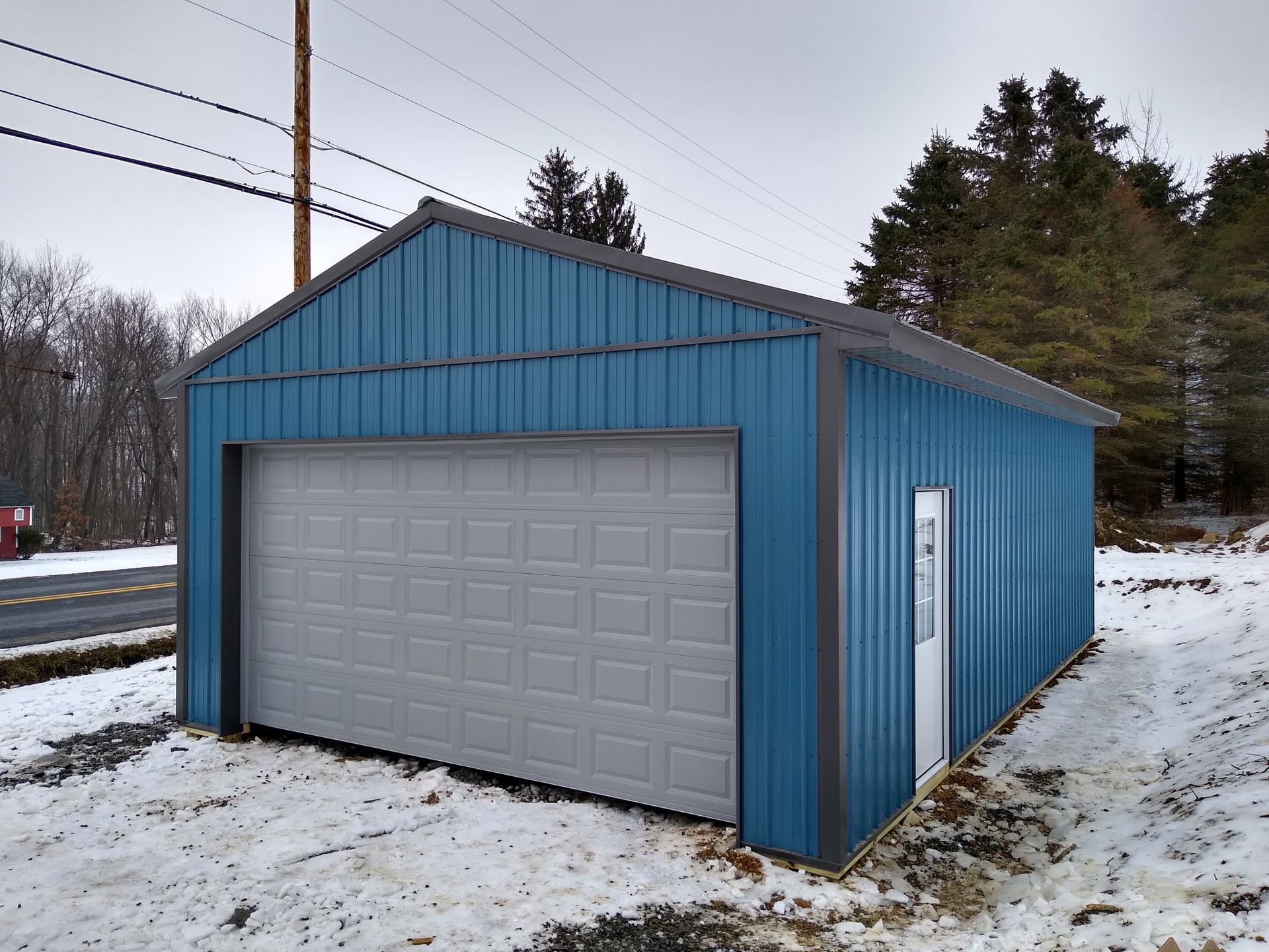 Blue metal garage with a gray door in a snowy yard, viewed from the front-left side.