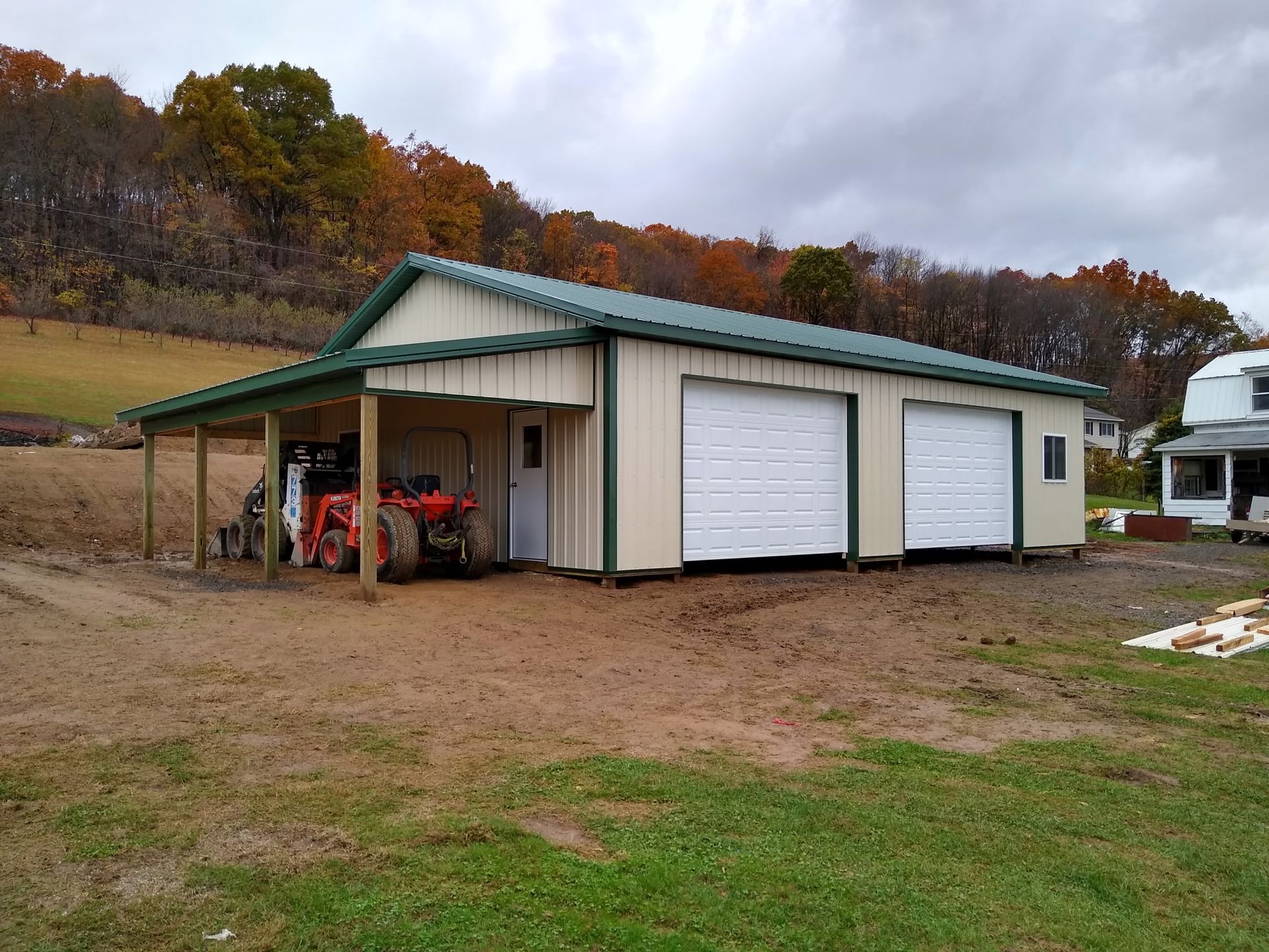 Rural metal garage with green roof and white doors beside a dirt yard and autumn trees