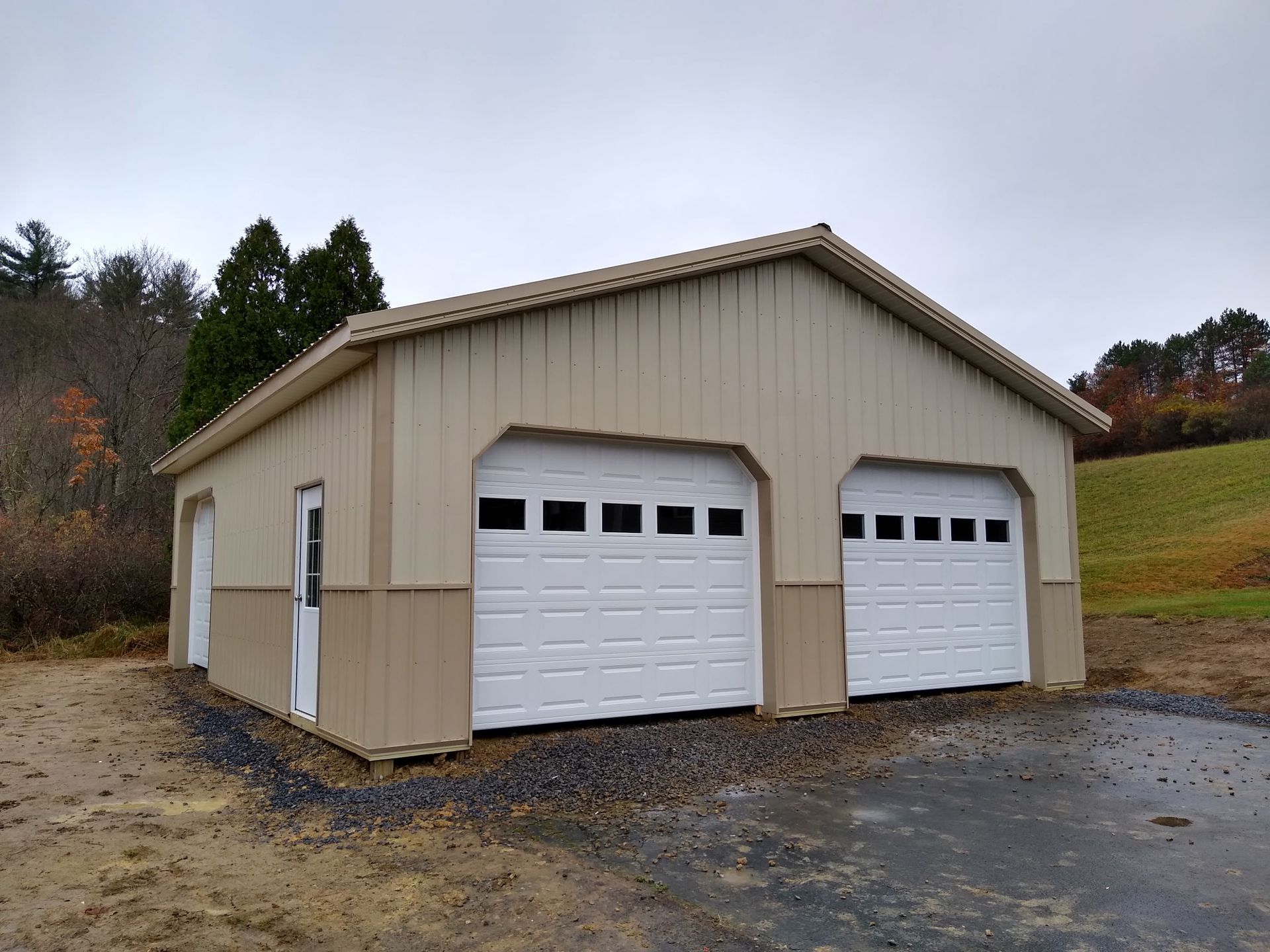 Beige two-car garage with white doors on a gravel driveway, surrounded by trees and a grassy slope.