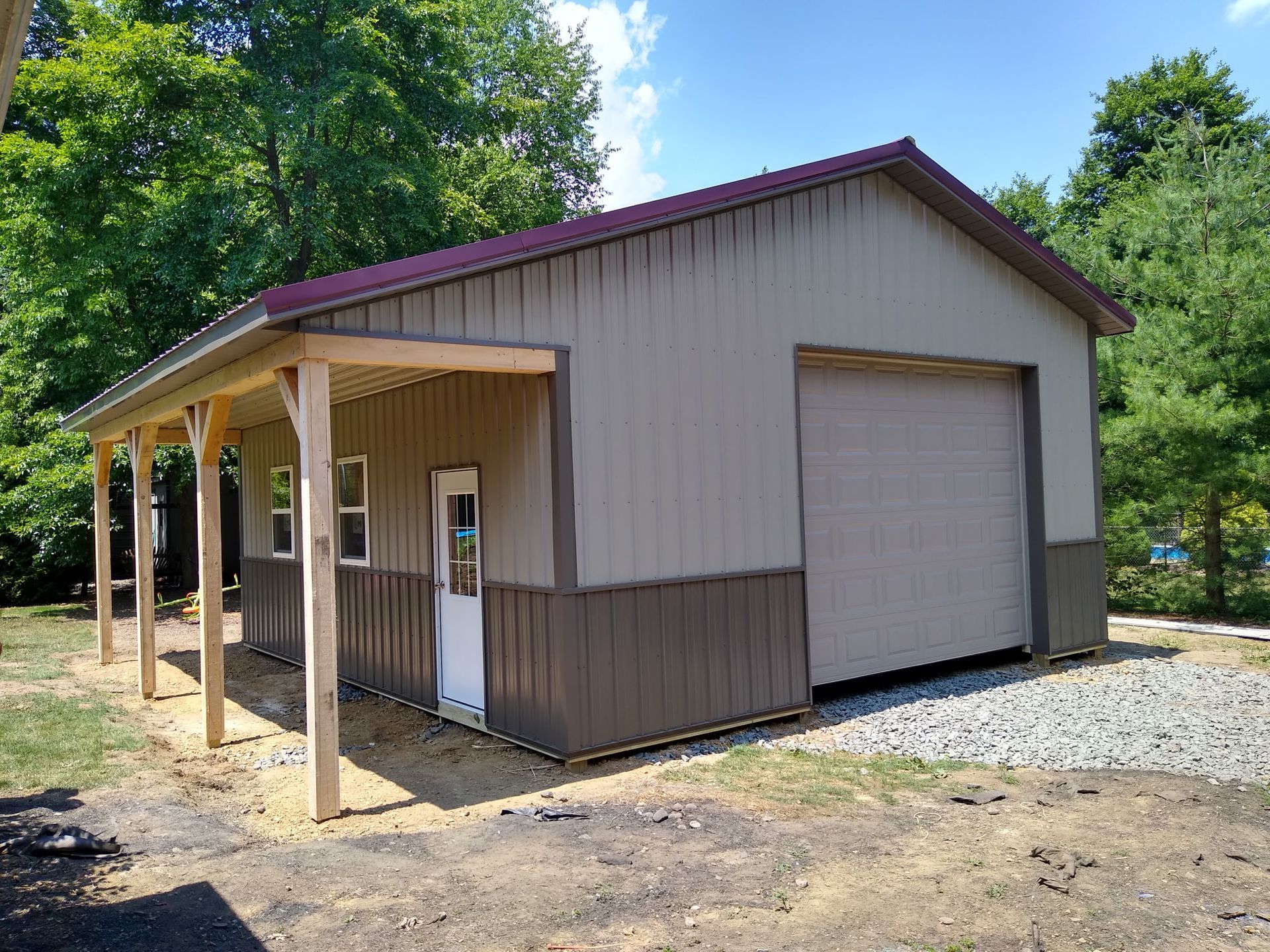 Tan metal-sided building with porch, roll-up door, and gravel lot, surrounded by trees