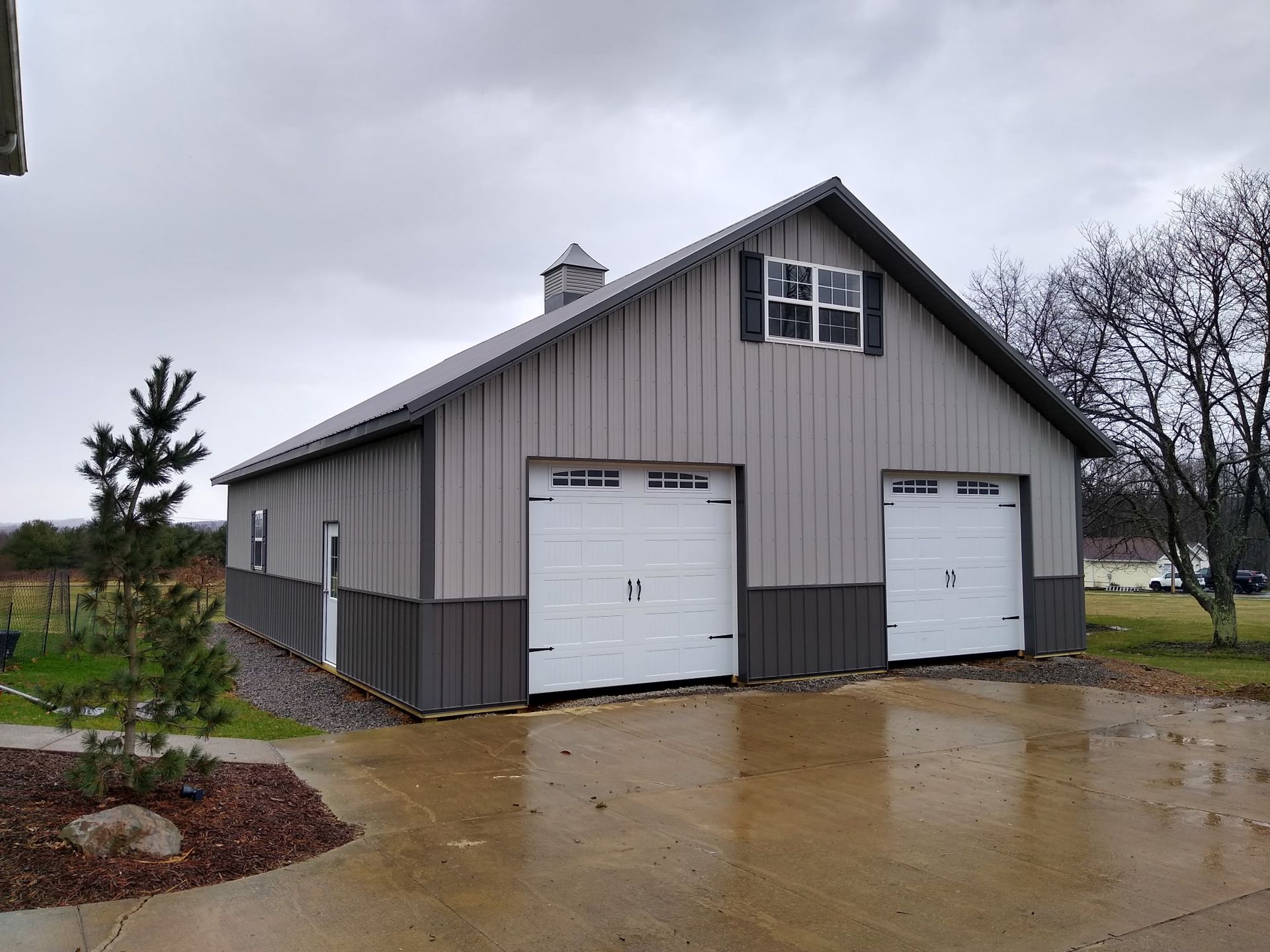 Gray barn-style garage with two white doors on a wet driveway in a rural setting