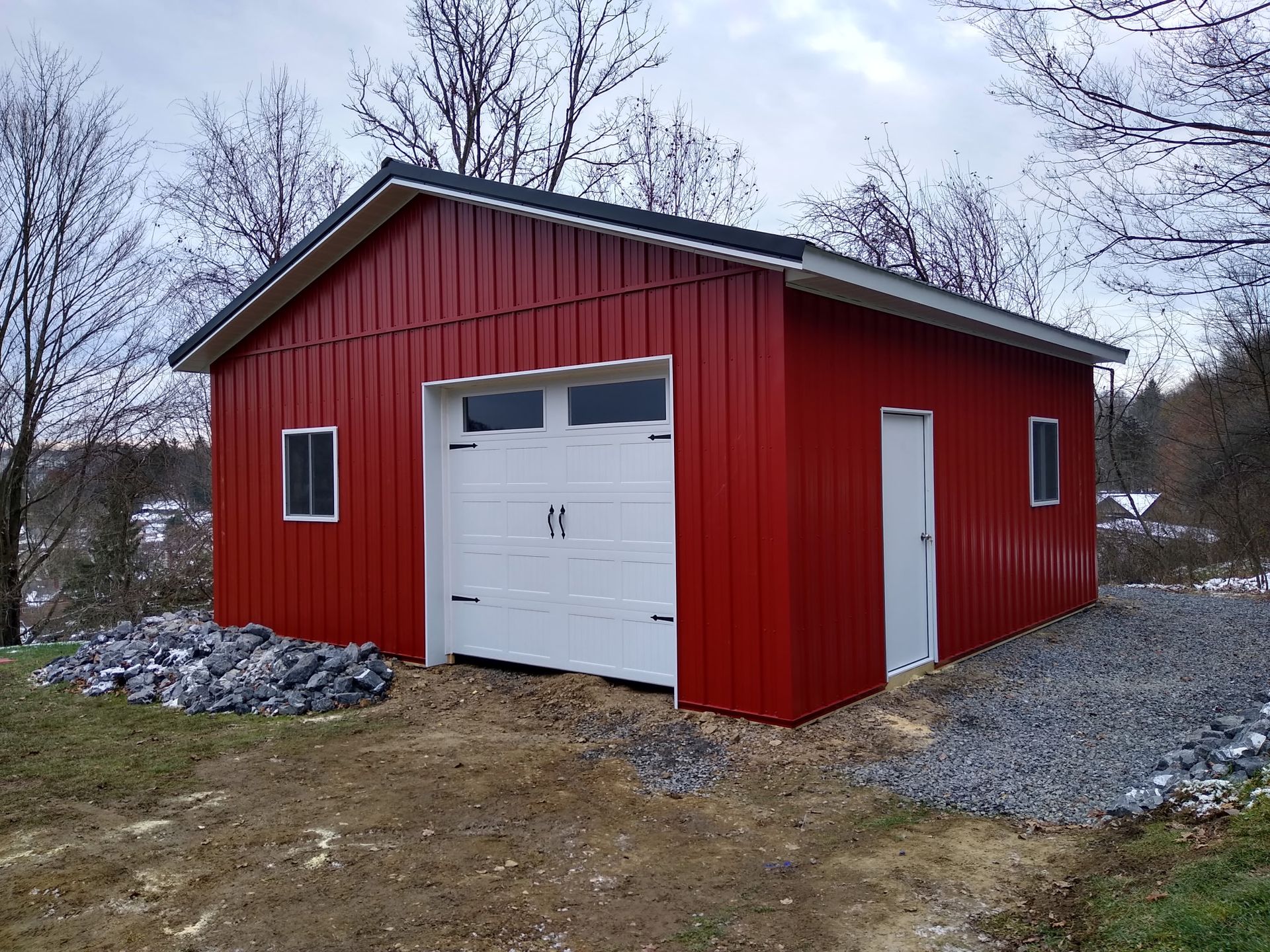 Red barn-style garage with white doors on a gravel lot in a leafless rural setting