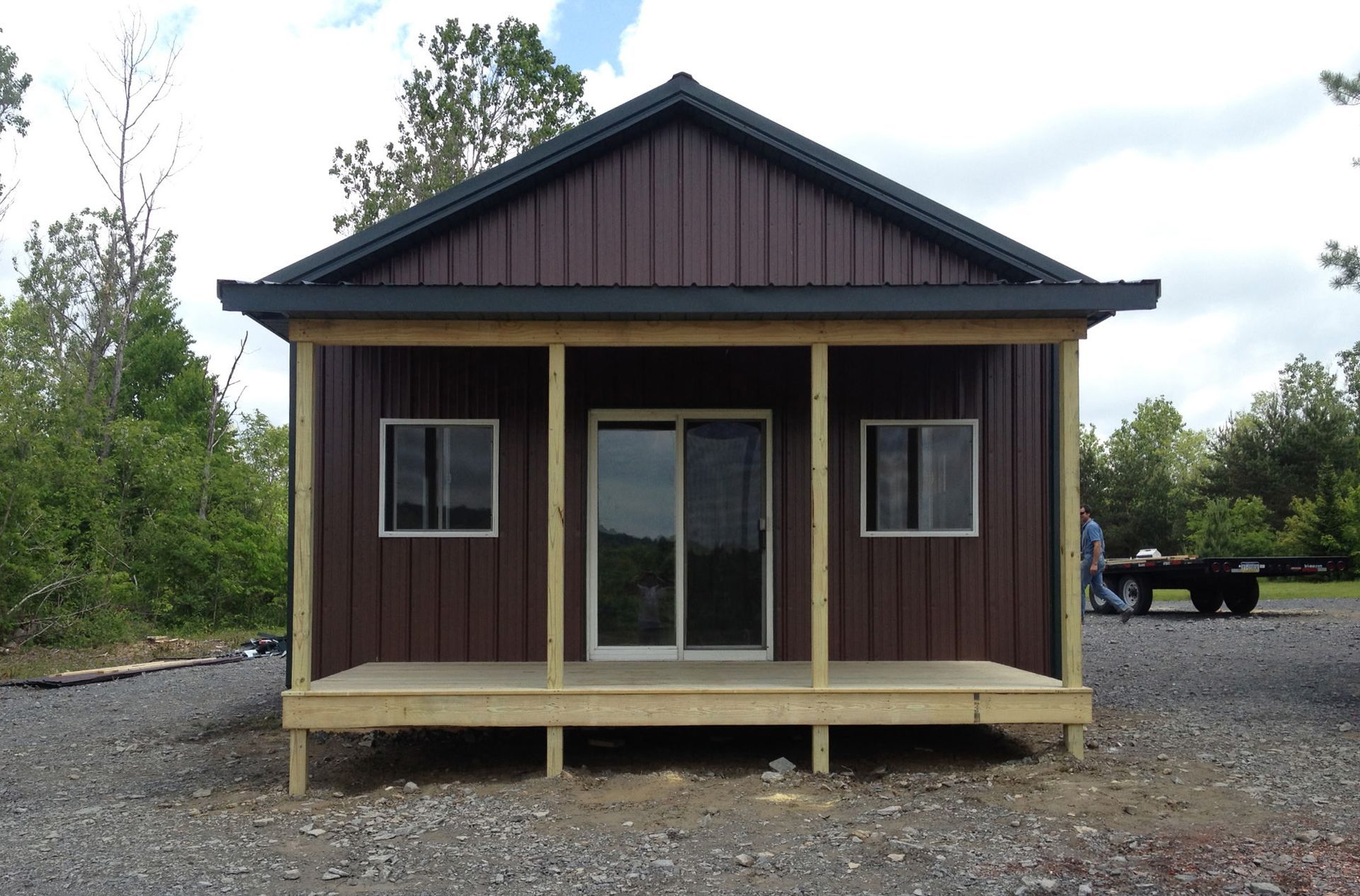 Small brown cabin with a front porch and double glass doors, set on a gravel lot with trees behind it