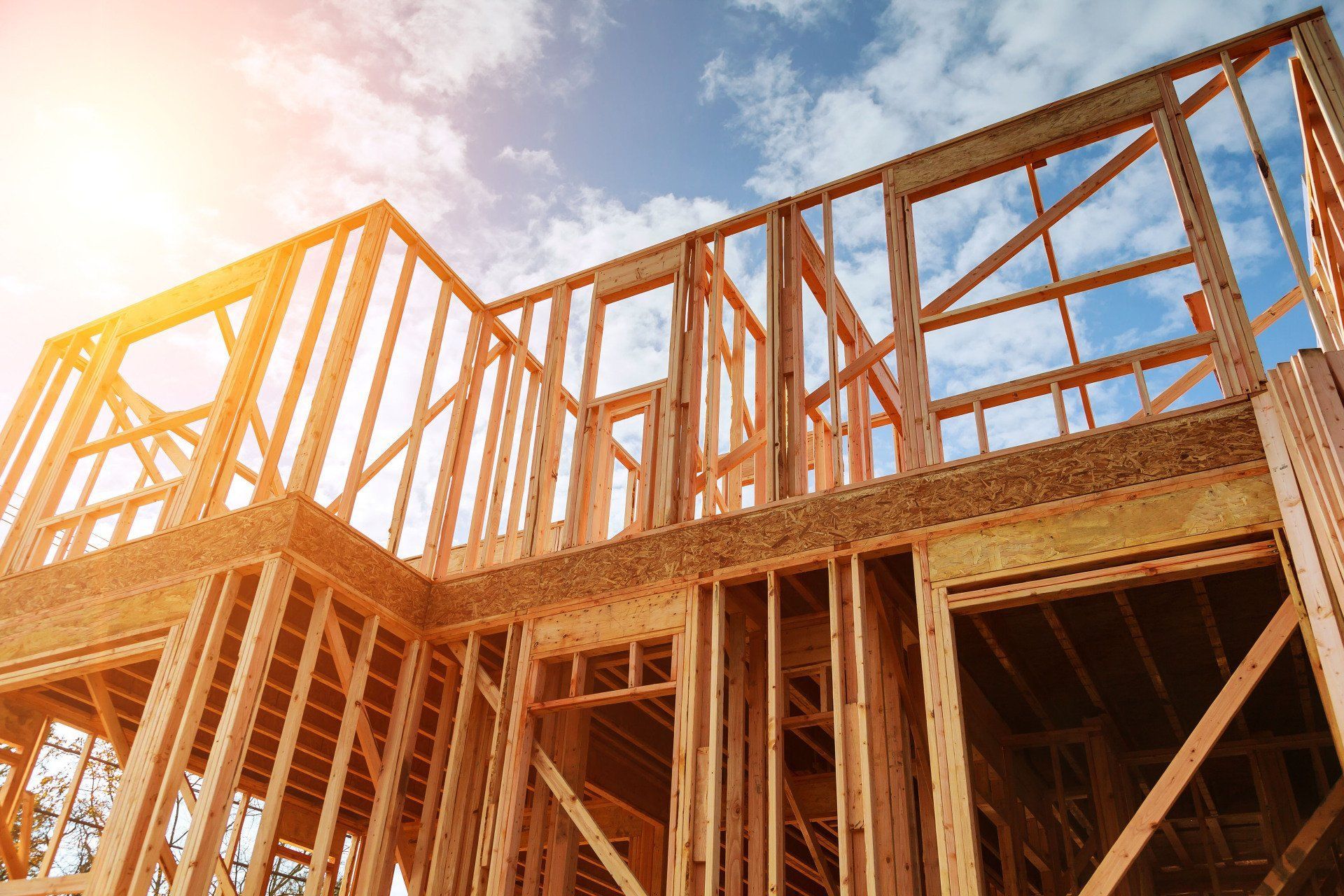 Wooden frame of a house under construction, with a bright sun and blue sky in the background.