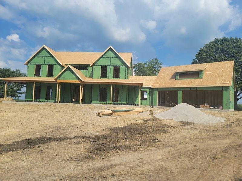 House under construction with green siding, brown roof, and garage on a dirt lot under a cloudy sky.