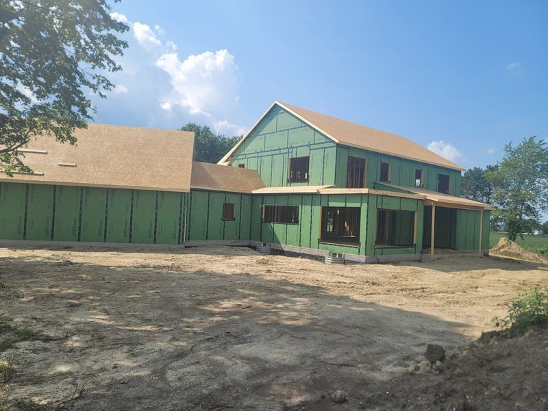 A two-story house under construction with green sheathing and brown roofing on a sunny day.