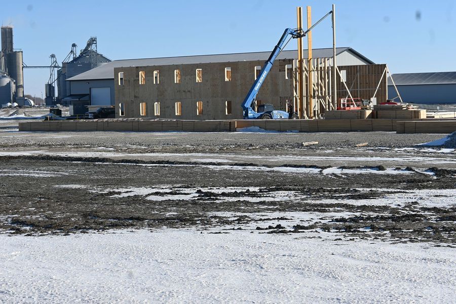 Construction site: wooden structure, blue lift, grain silos in background, snowy ground.