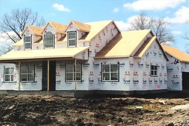 A house under construction, wrapped in Tyvek. Front porch, dormers, and windows visible.