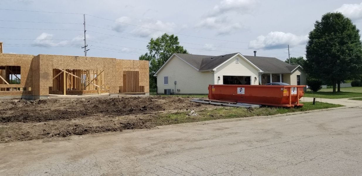 Construction site with a partially built wooden structure and a small white house with a dumpster in front.