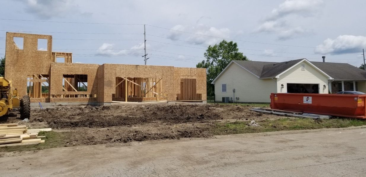 A building under construction with exposed wooden frame. A finished house and dumpster are nearby. Cloudy sky.