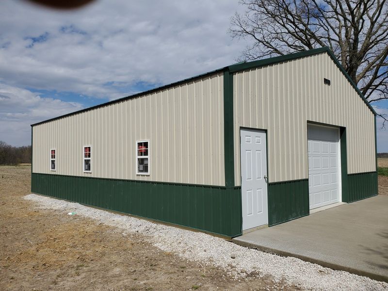 Tan and green metal building with a white door, garage door, and windows, on a concrete pad.