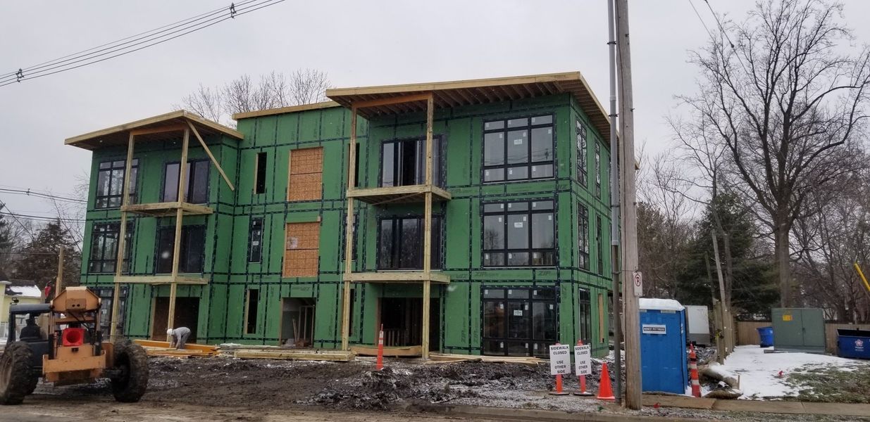 Multi-story building under construction with green siding, large windows, and balconies. An excavator is in the foreground.