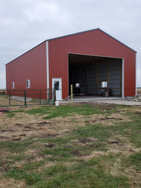 Red metal barn with large opening, white trim, and small windows on a grassy field under a cloudy sky.