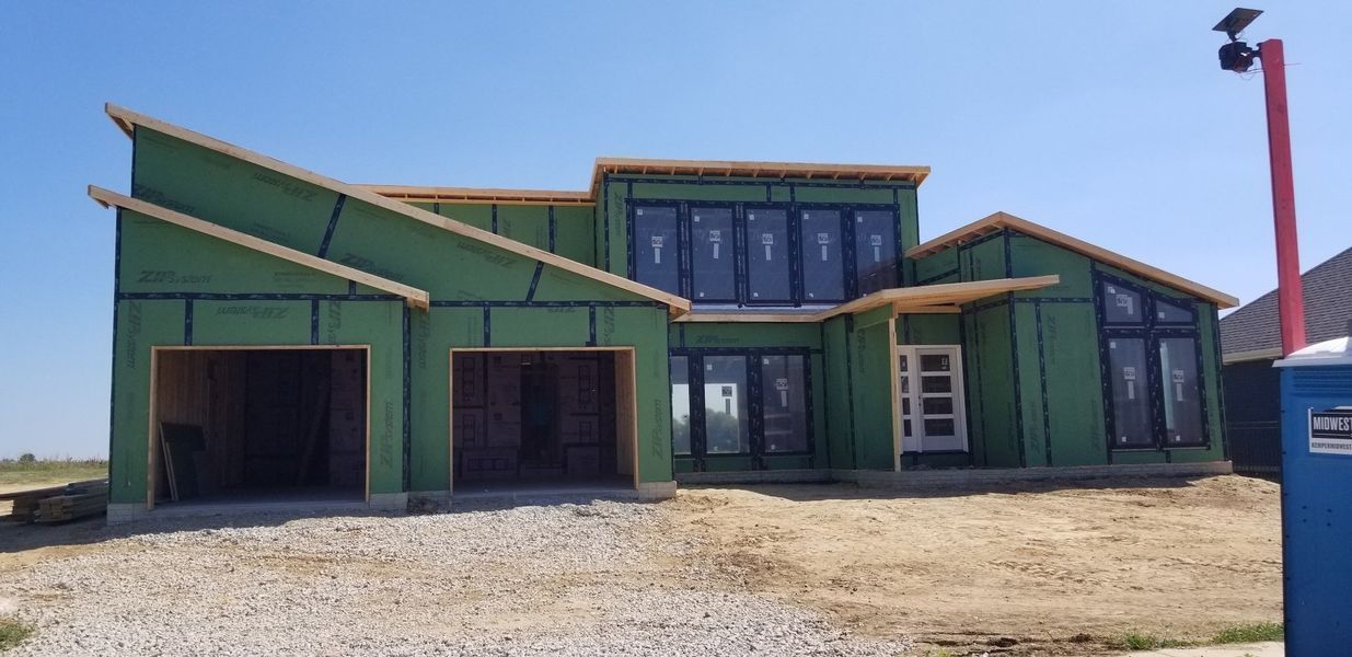 House under construction, green sheathing, two-car garage, large windows, blue portable toilet to the right.