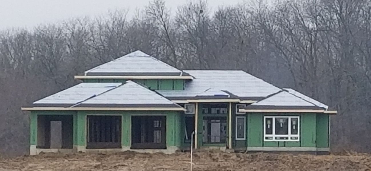 A house under construction with a snow-covered roof, surrounded by bare trees under an overcast sky.