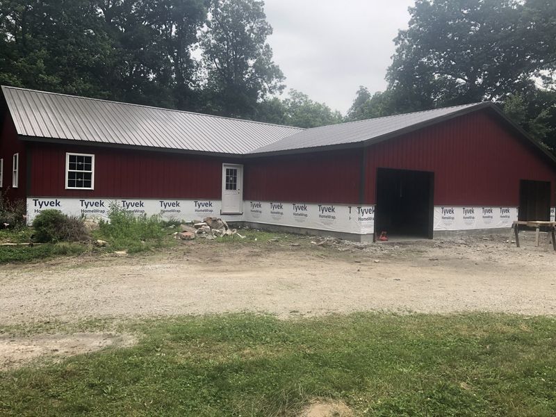 Red metal-roofed building with white trim, a door, and a garage door, set on a gravel driveway, surrounded by trees.