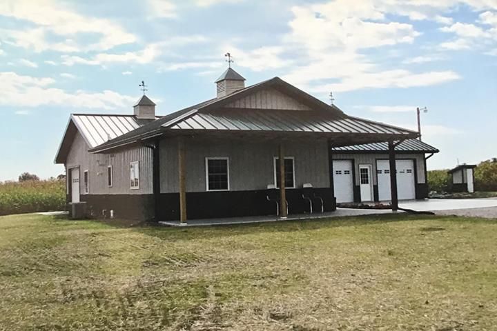 Gray and brown barn-style building with a covered porch and garage bays; field in the foreground, cloudy sky.