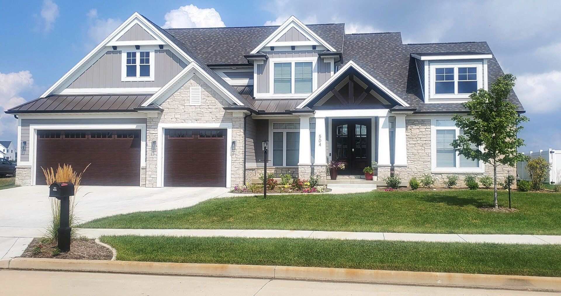 Two-story house with gray siding, stone accents, brown garage doors, and a well-manicured lawn under a blue sky.