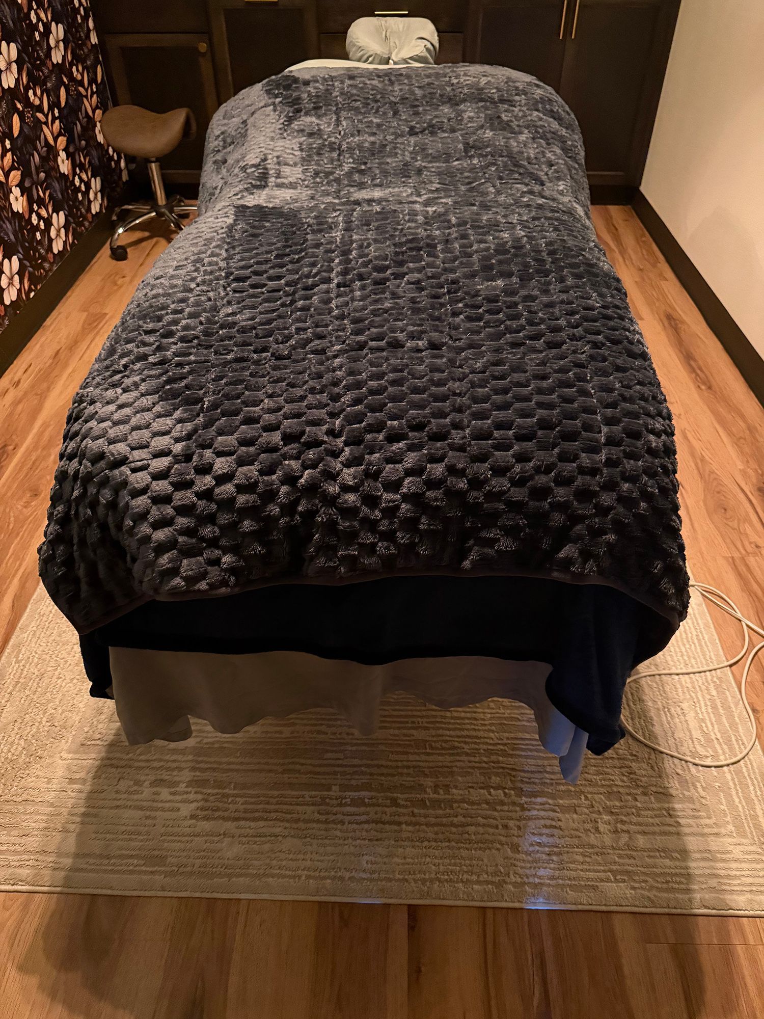 A bed with a dark textured blanket in a warmly lit spa room, viewed from the foot of the bed.