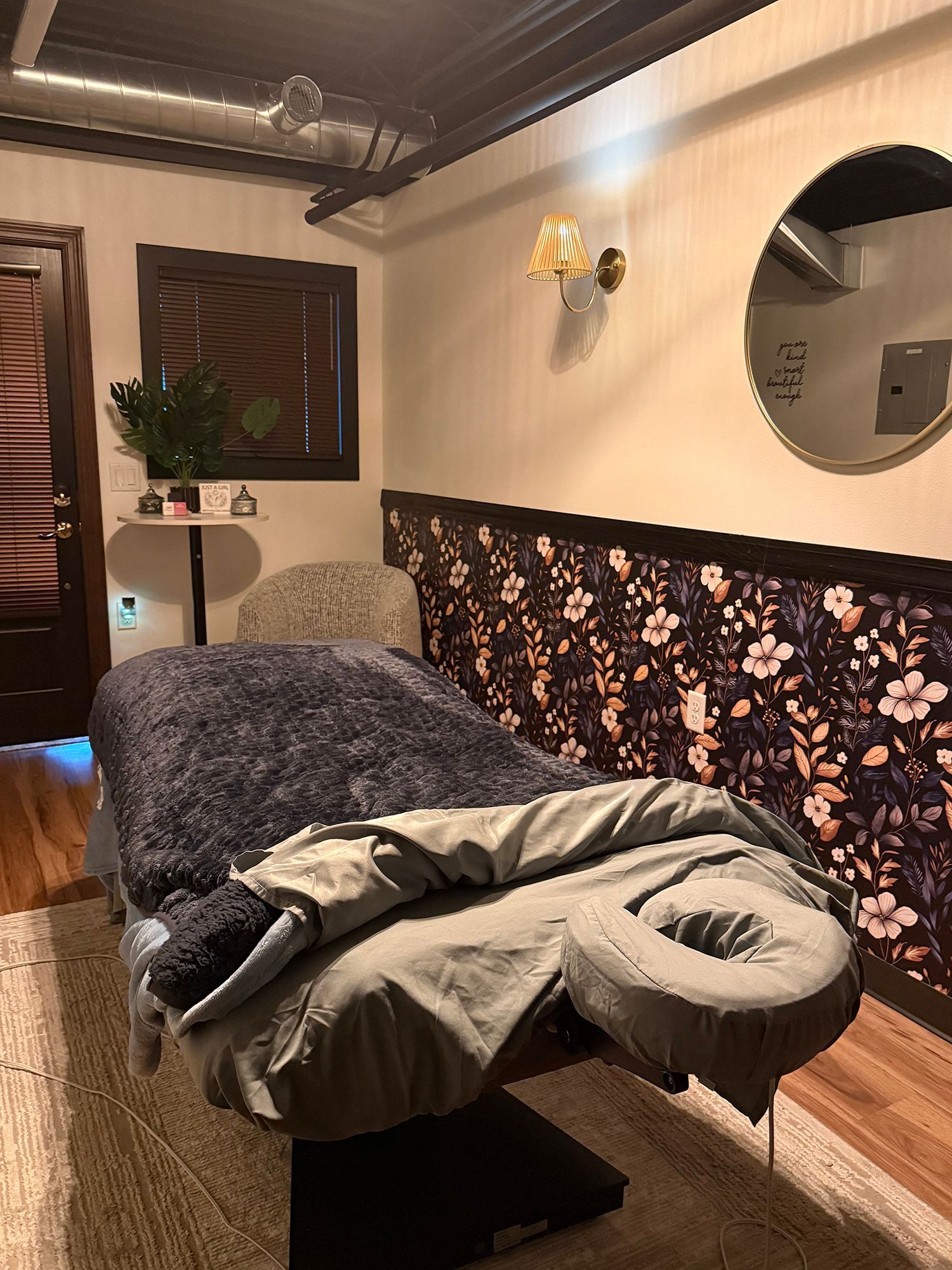 Massage table in a softly lit spa room with floral wallpaper, round mirror, and wooden floor.