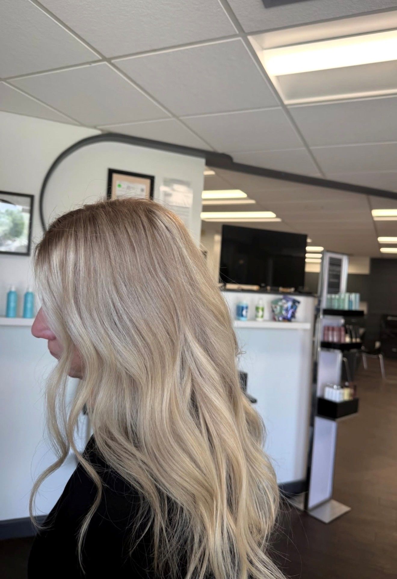 Woman with long blonde hair in a salon interior, seen in profile near a mirror and reception area