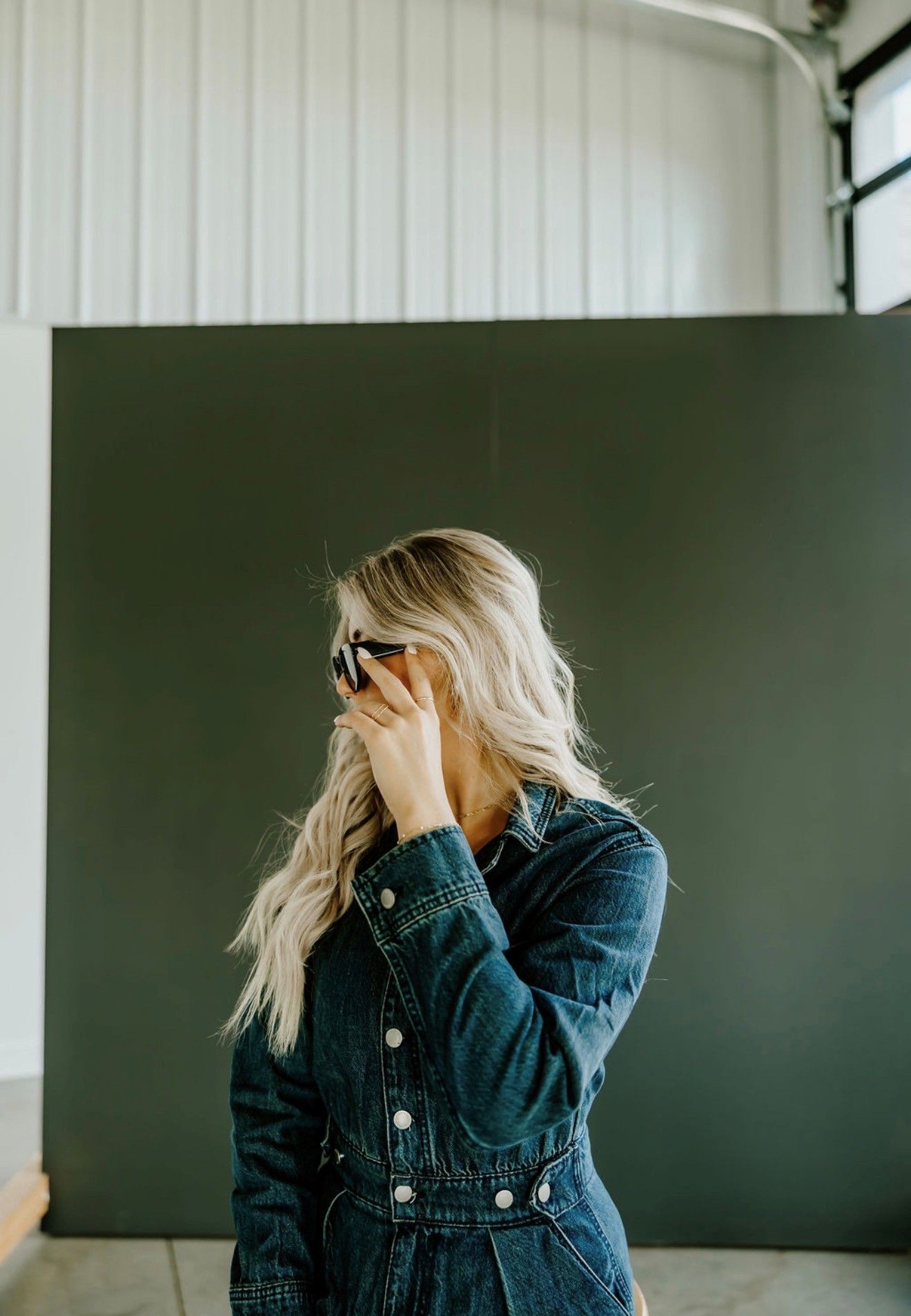 Woman in denim jacket adjusting sunglasses before a dark green backdrop