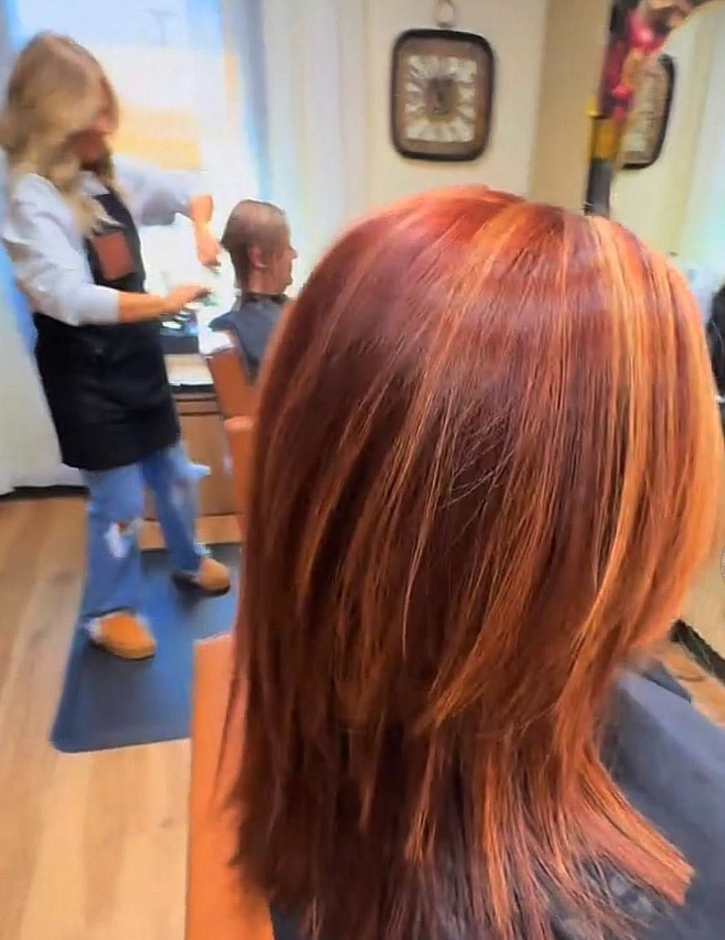 Red-haired person in a salon chair, with a stylist and seated child in the background.