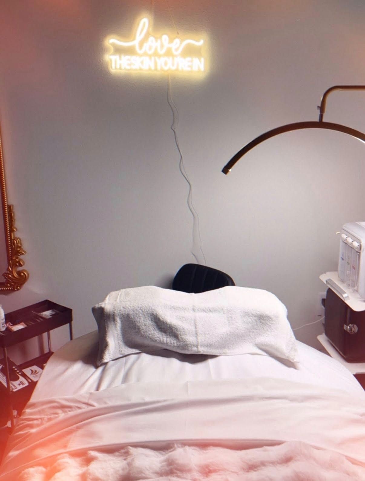 A cozy bedroom with a neon sign, white bed, black chair, and warm ambient lighting.