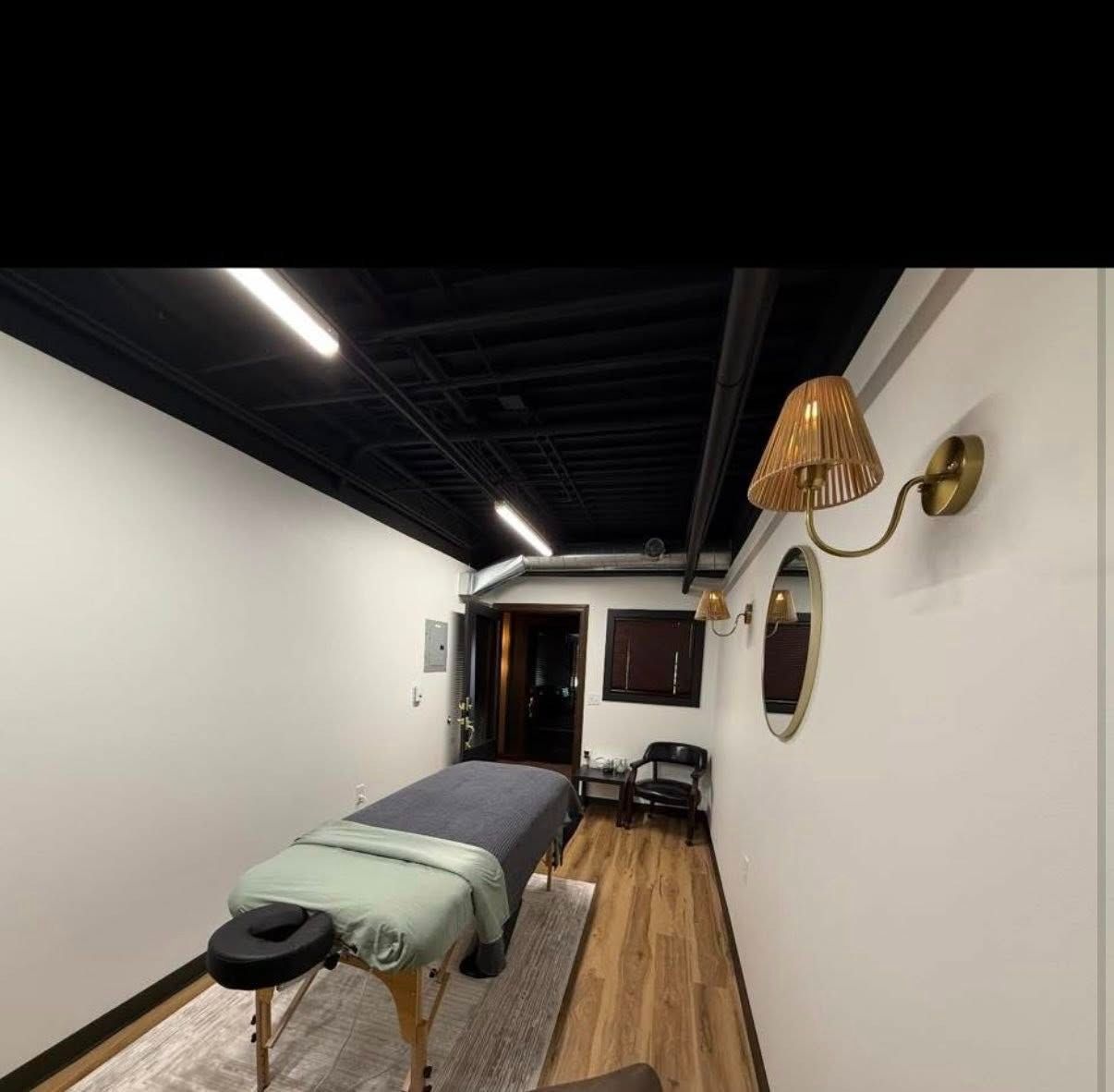 Massage therapy room with a treatment table, wood floor, white walls, and a woven wall sconce.