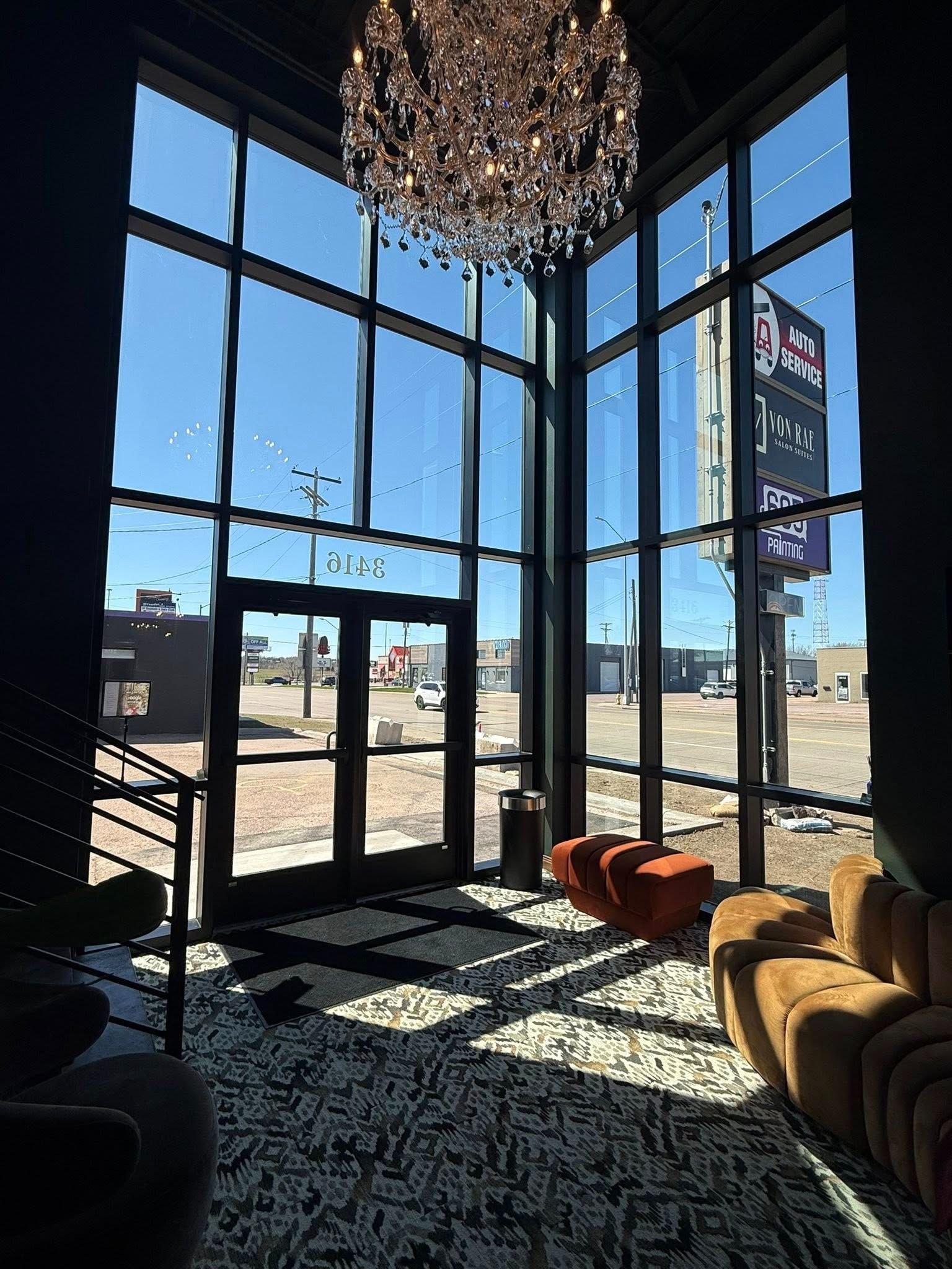 Modern lobby with floor-to-ceiling windows, chandelier, patterned rug, and orange chairs overlooking a street.