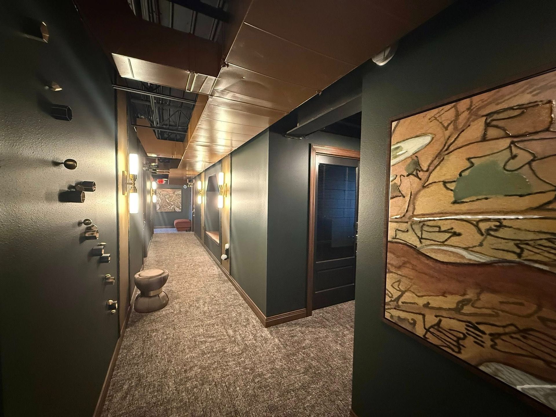 Dim hotel hallway with patterned carpet, dark walls, and warm ceiling lights leading to a bright end.