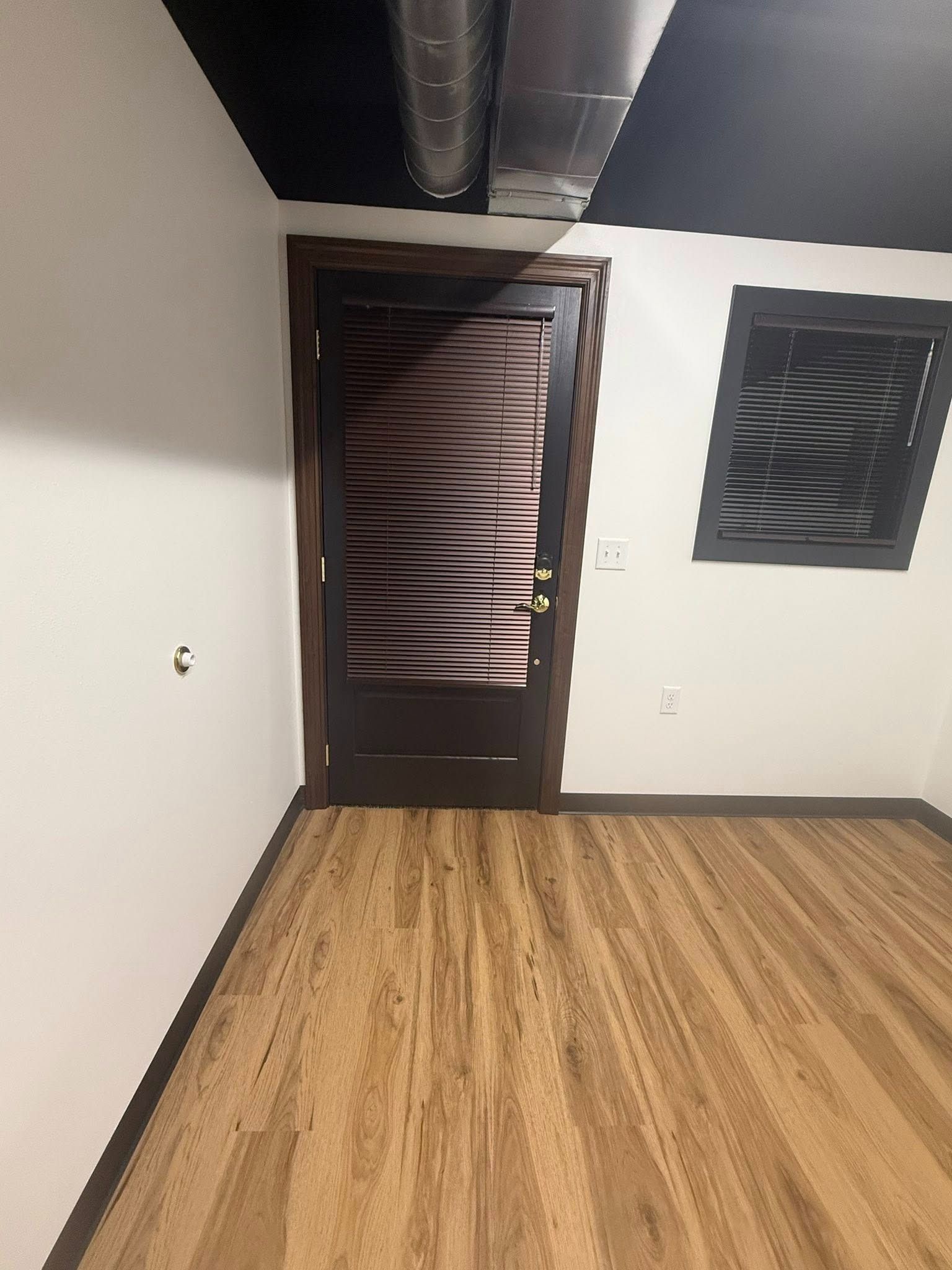 Empty room with wood flooring, dark door, white walls, and exposed ceiling ductwork.