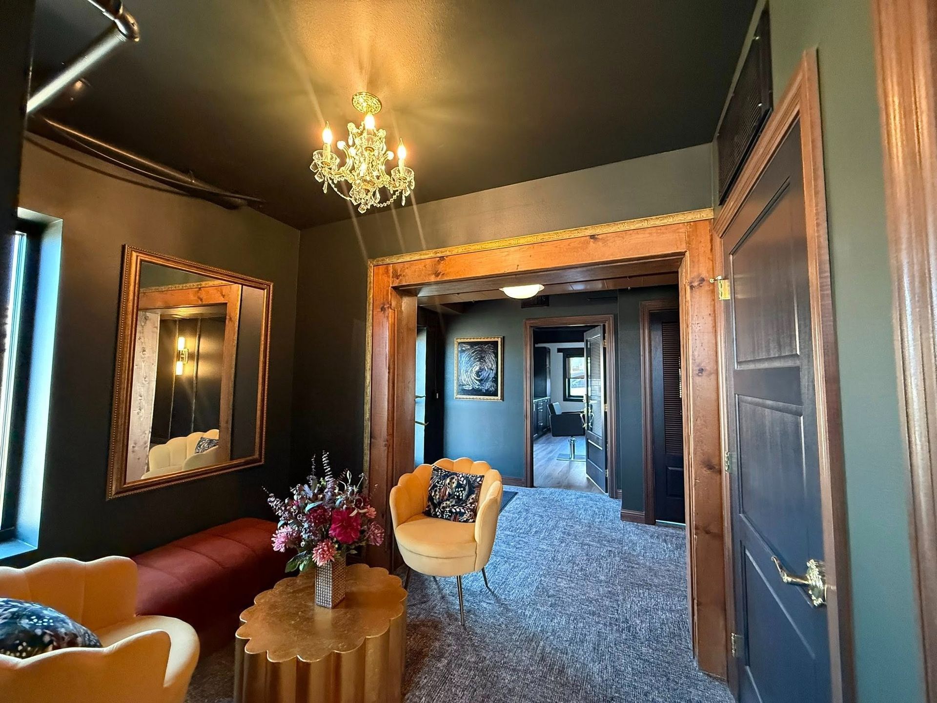 Elegant dark hallway with chandelier, framed mirror, upholstered chairs, and floral arrangement