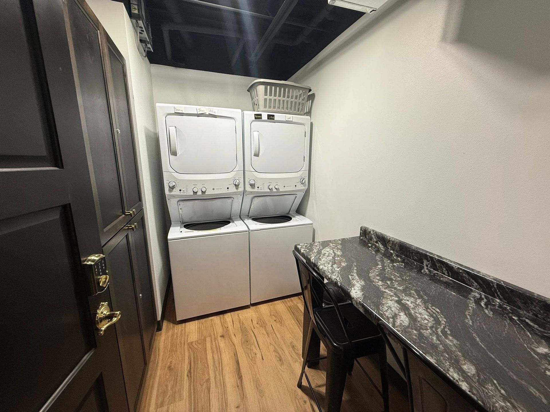 Laundry room with stacked white washers and dryers beside a black counter and chair on wood flooring