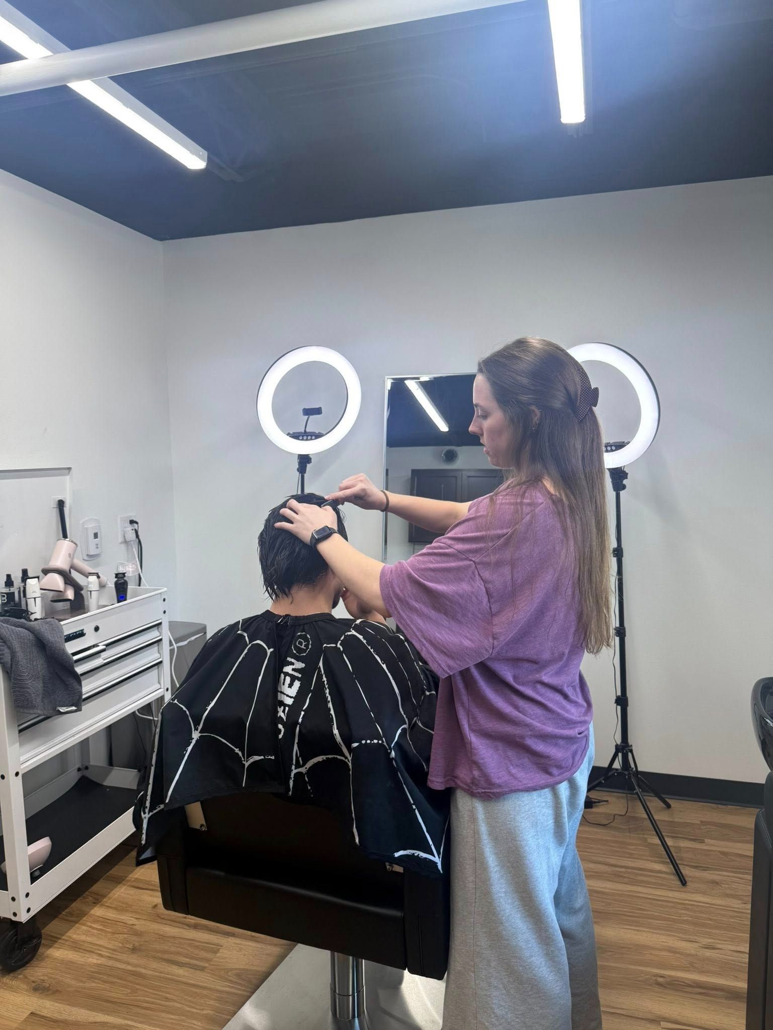 Stylist trimming a client’s hair in a modern salon with ring lights and a mirror.